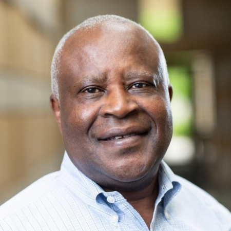 A middle-aged African American man with a bald head, wearing a light blue collared shirt, smiling in an indoor setting with blurred background.