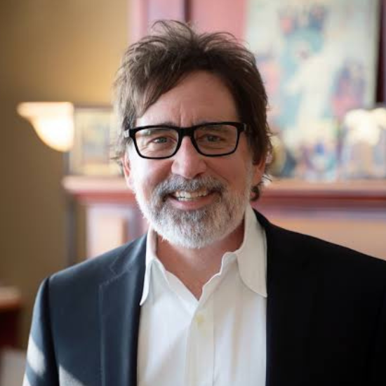 A middle-aged man with glasses, gray hair, and a beard, smiling, wearing a dark blazer and a white shirt, standing indoors with a blurred background.