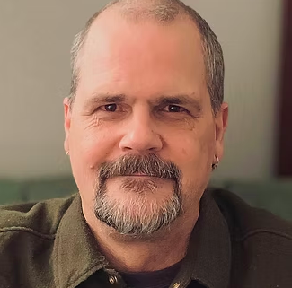 Close-up of a middle-aged man with short gray hair, beard, and mustache, wearing a dark green shirt, sitting on a green couch in an indoor setting.