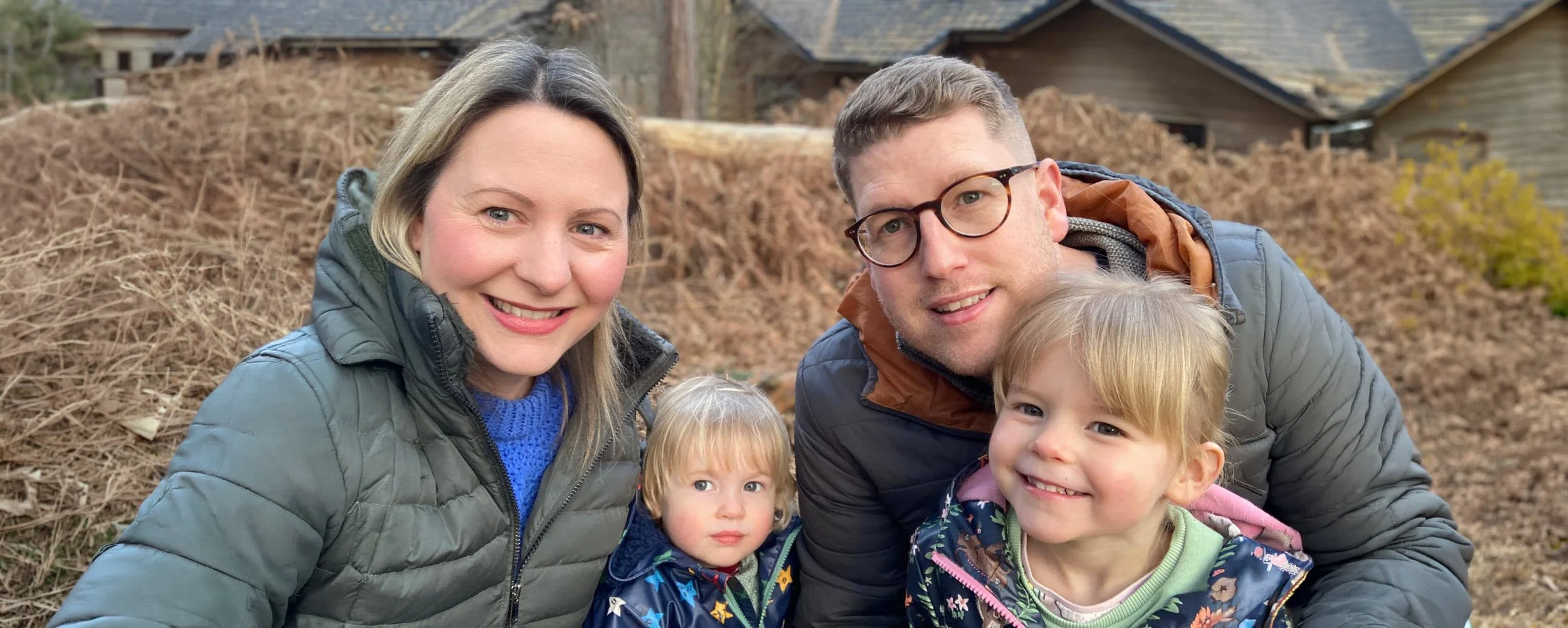 A family of four smiling outdoors, with two children, in front of a background of dried bushes and houses.