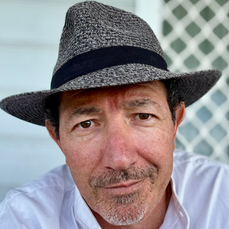 Close-up of a man with a slight smile wearing a gray fedora hat and a white shirt, with a lattice fence in the background.