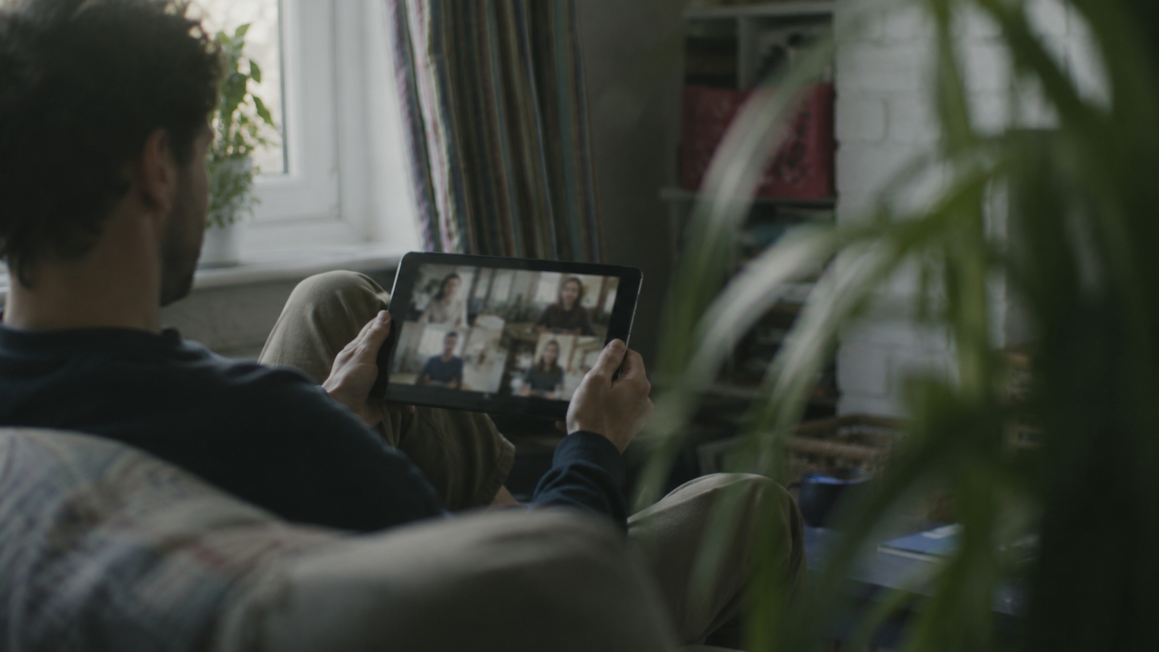 A person sitting on a couch holding a tablet with a video call showing five people.