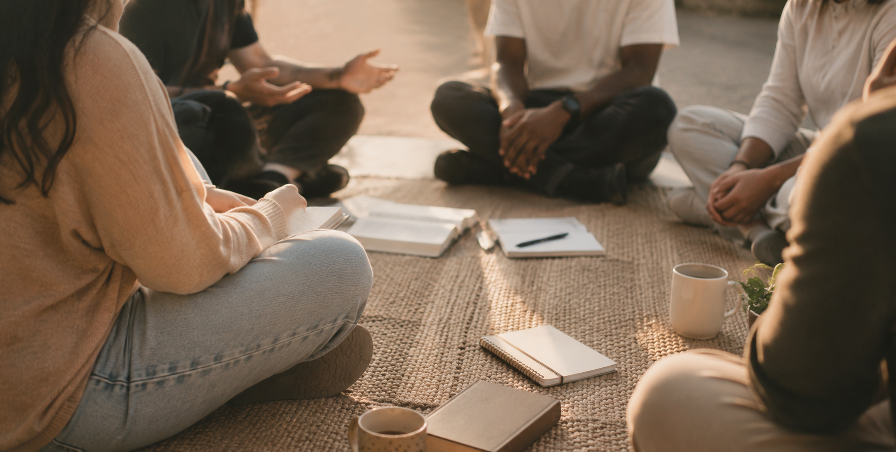 Group of people sitting on the floor in a circle with notebooks, pens, and coffee mugs in a cozy setting.
