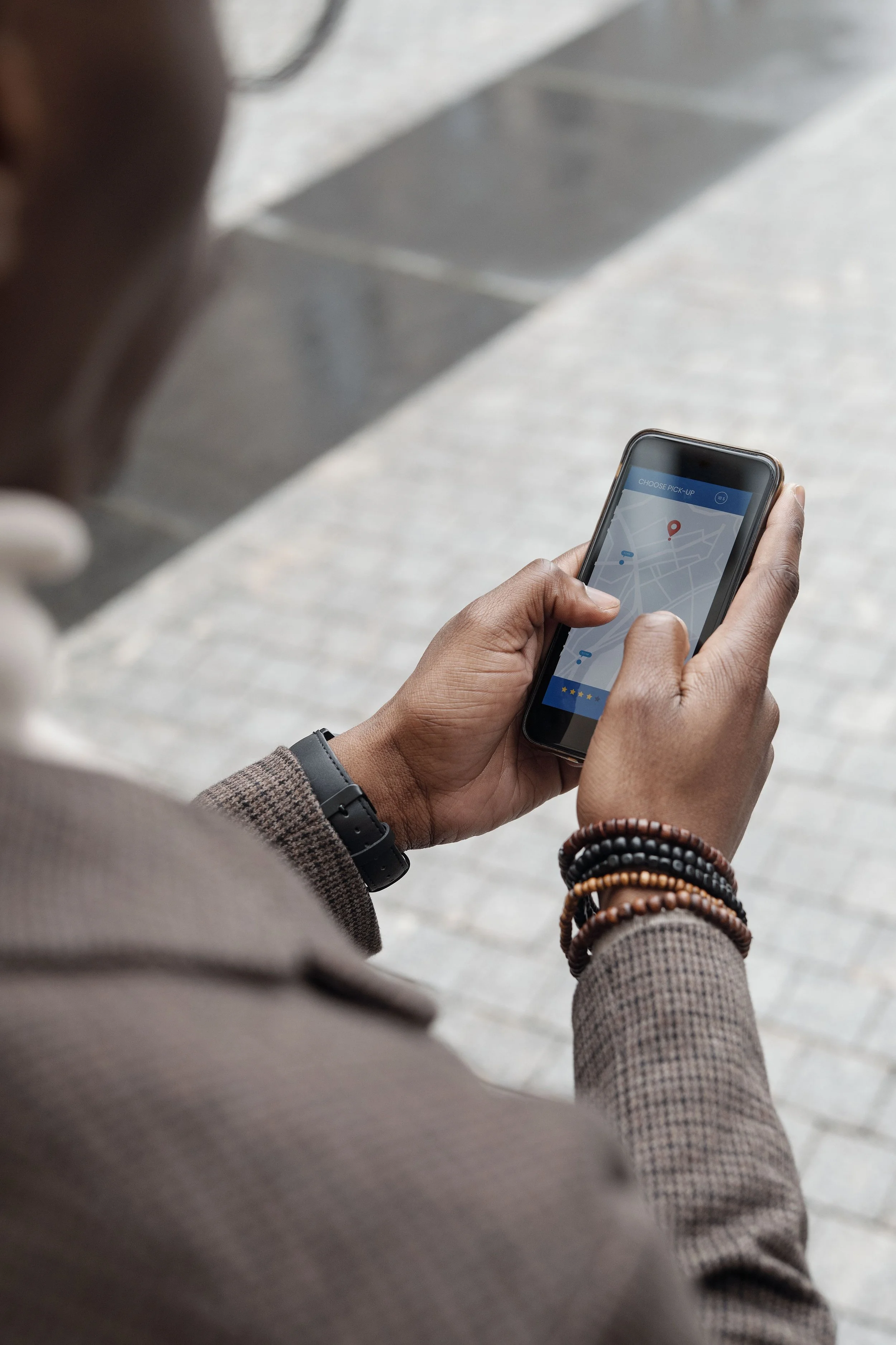 Person holding smartphone with a map app open, person wearing multiple beaded bracelets and a wristwatch, gray blazer, sidewalk in background.