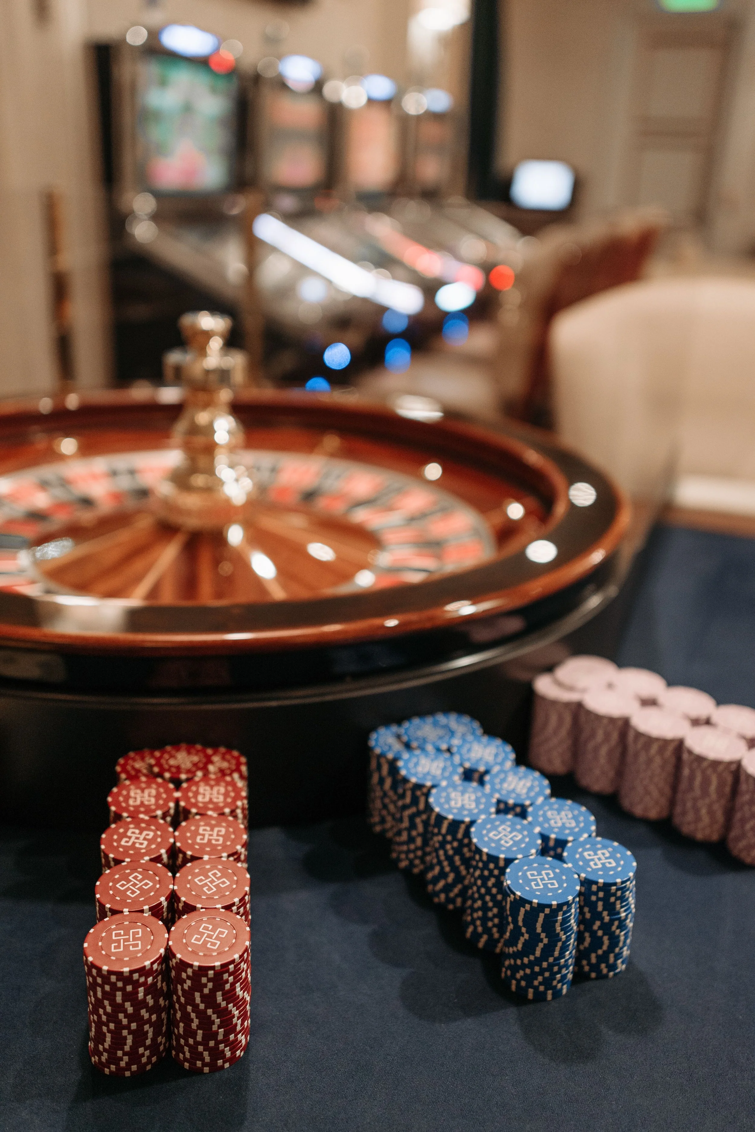 Stacks of poker chips in red, blue, and pink colors placed on a gaming table, with a roulette wheel and blurred gaming machines in the background.