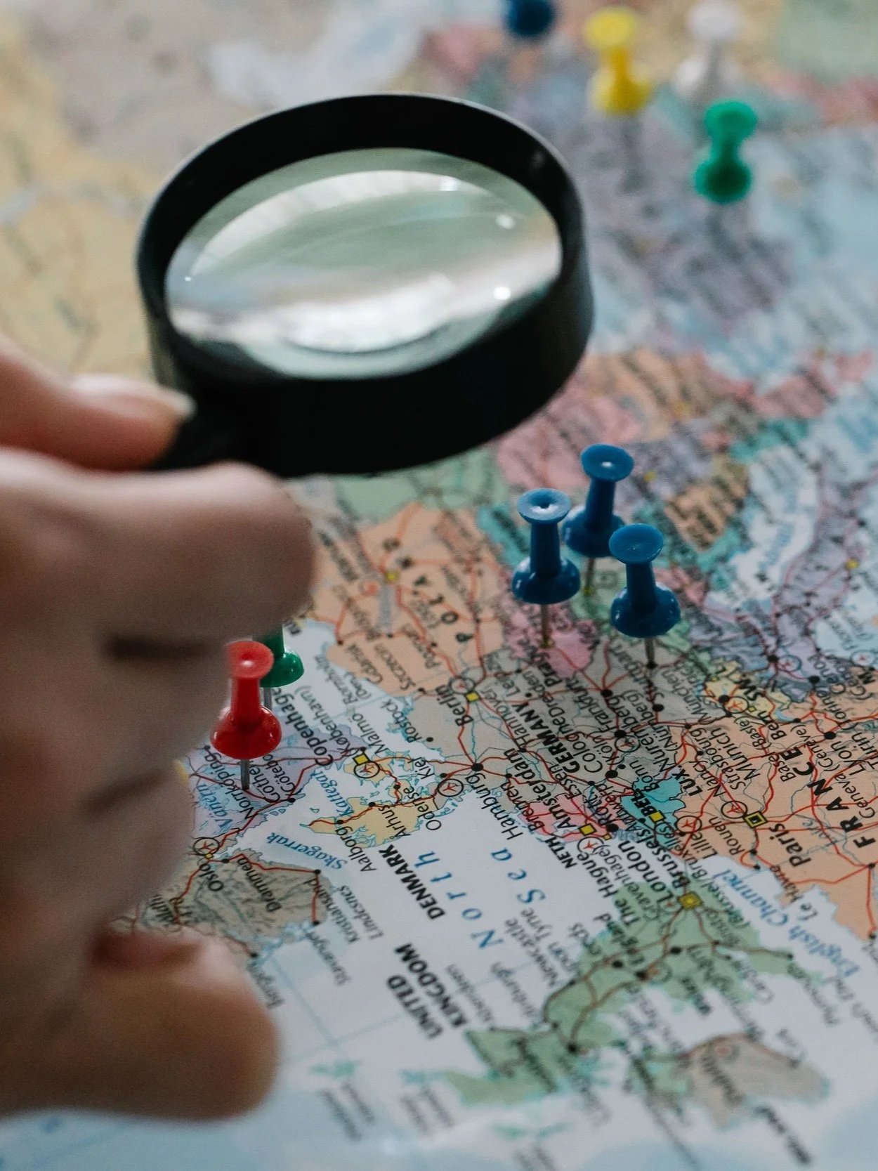 Person holding a magnifying glass over a colorful map with red, blue, yellow, and green push pins.