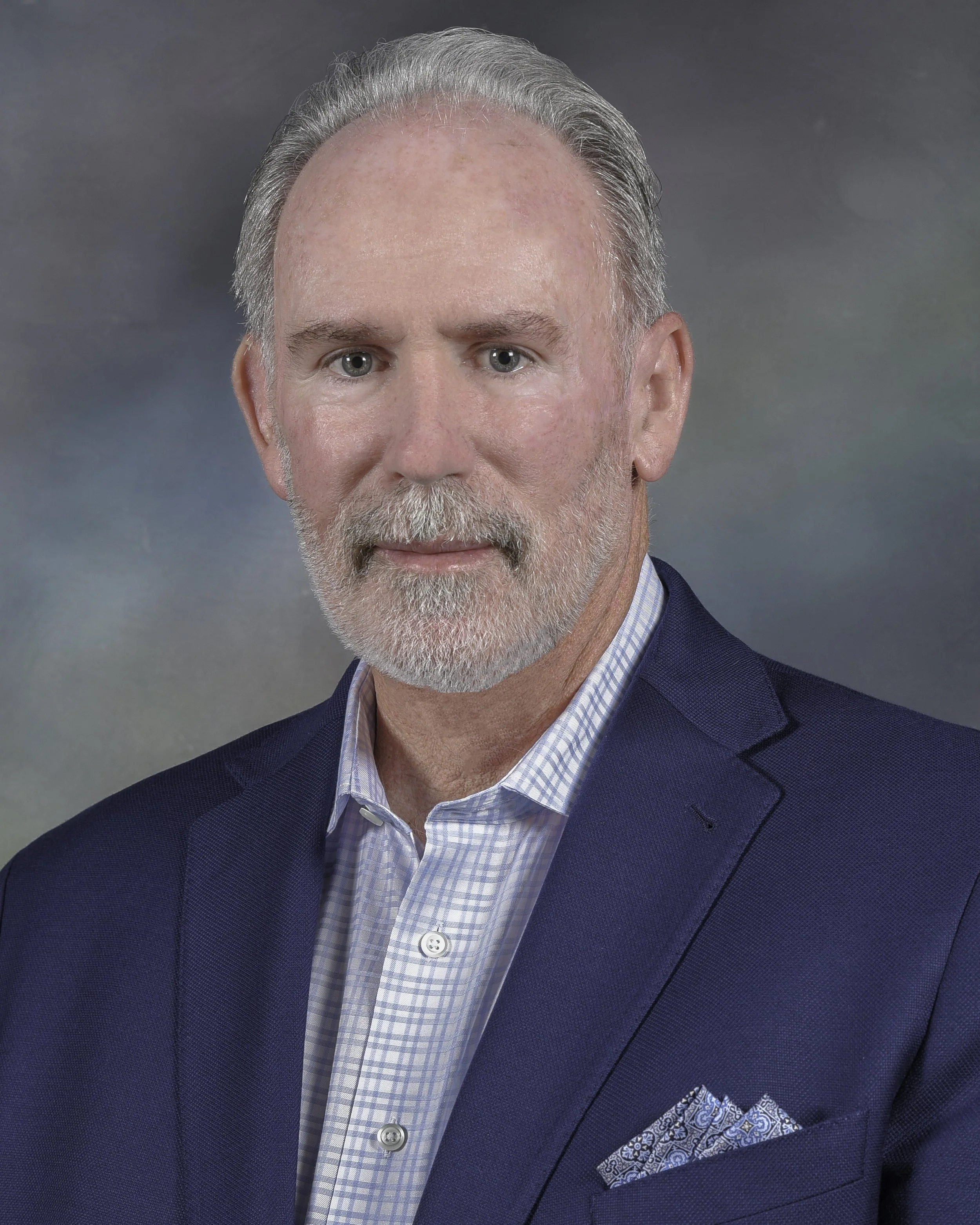 Close-up of a middle-aged man with gray hair and beard wearing a dark blue blazer, light blue checkered shirt, and a matching blue pocket square against a gray background.