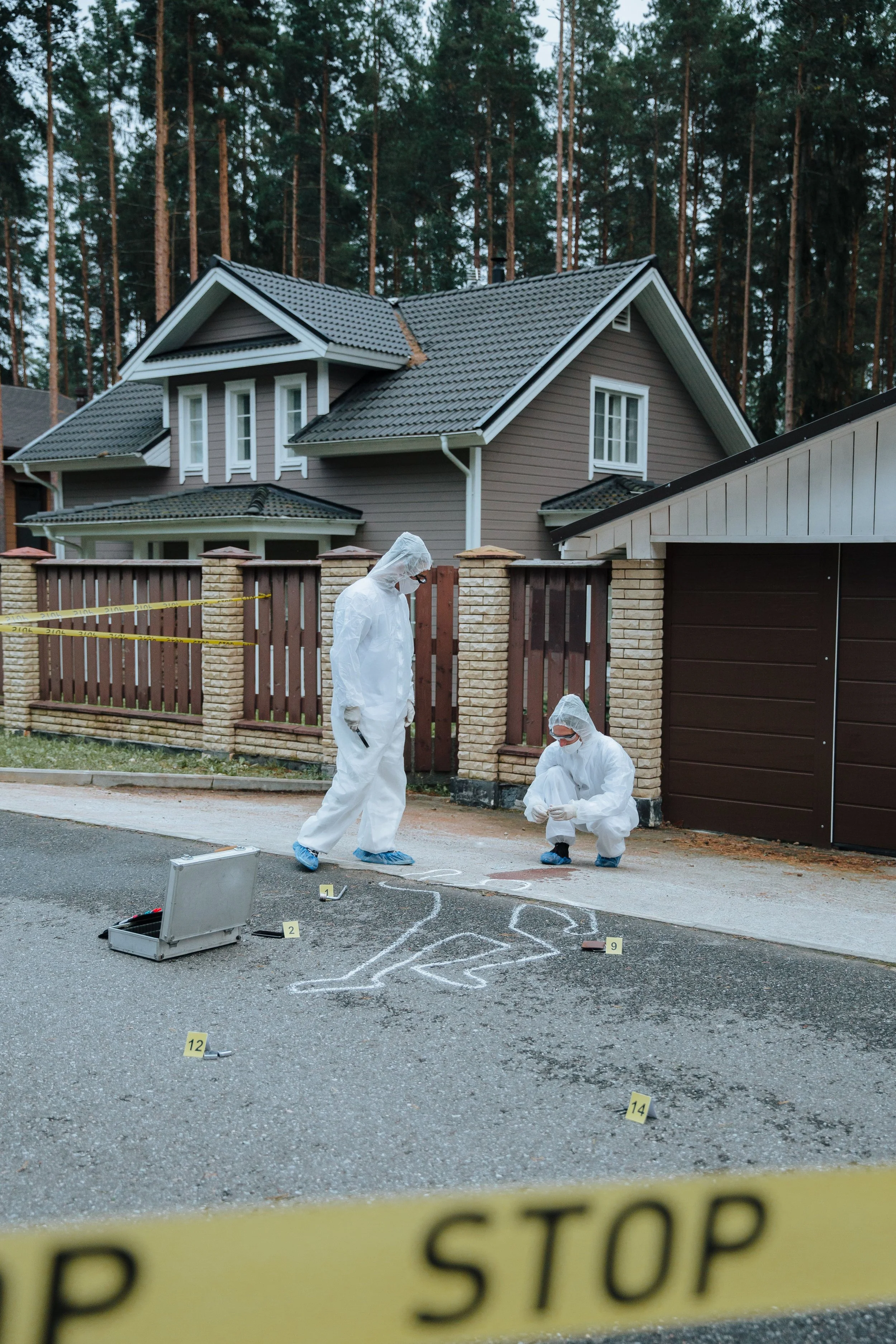 Forensic investigators analyzing a crime scene outdoors in front of a residential house with workers in protective white suits and masks. There are numbered evidence markers on the ground and yellow police tape in the foreground.
