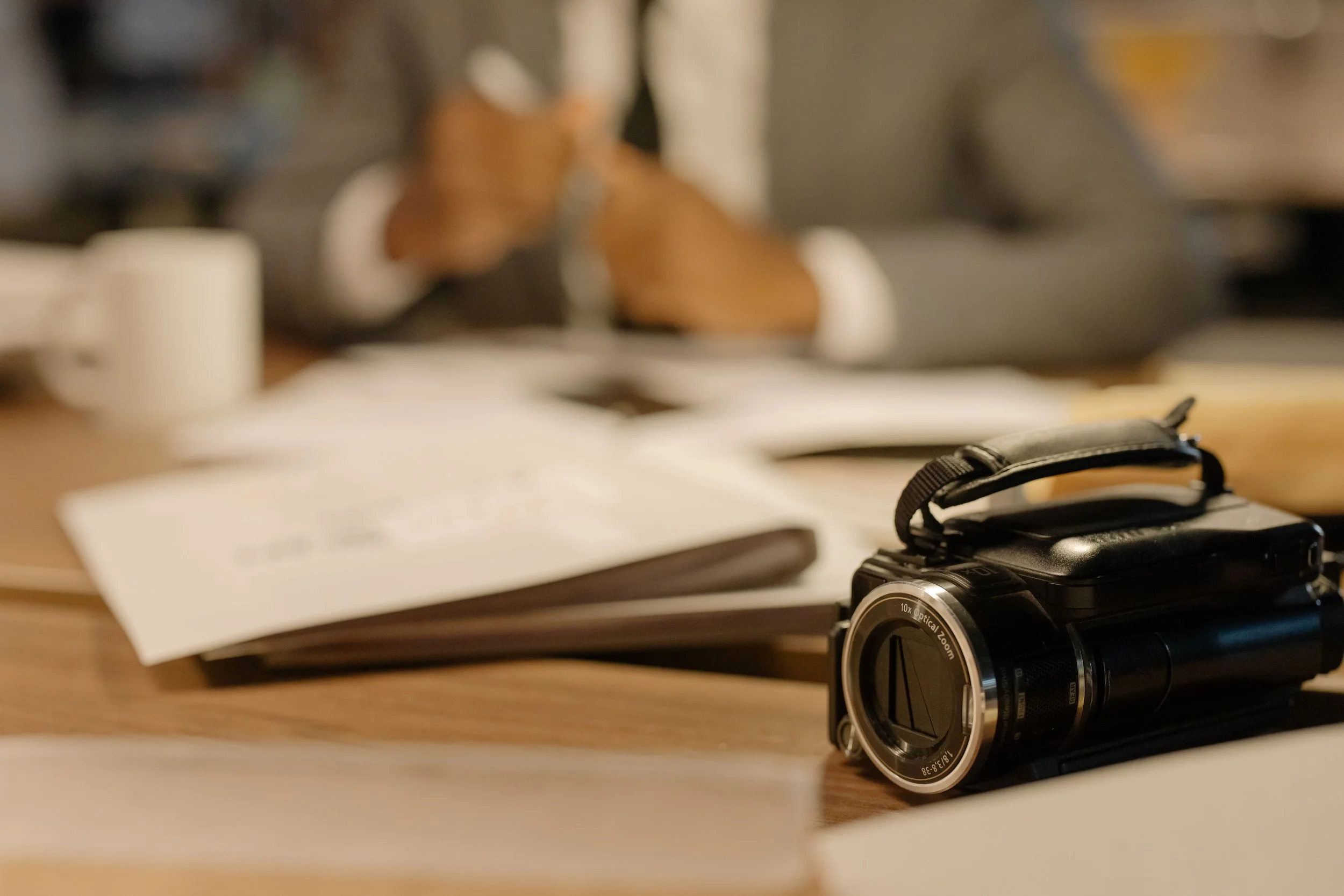 A black compact digital camera with a large lens on a wooden desk, with papers, a white mug, and a camera strap nearby, slightly out of focus.