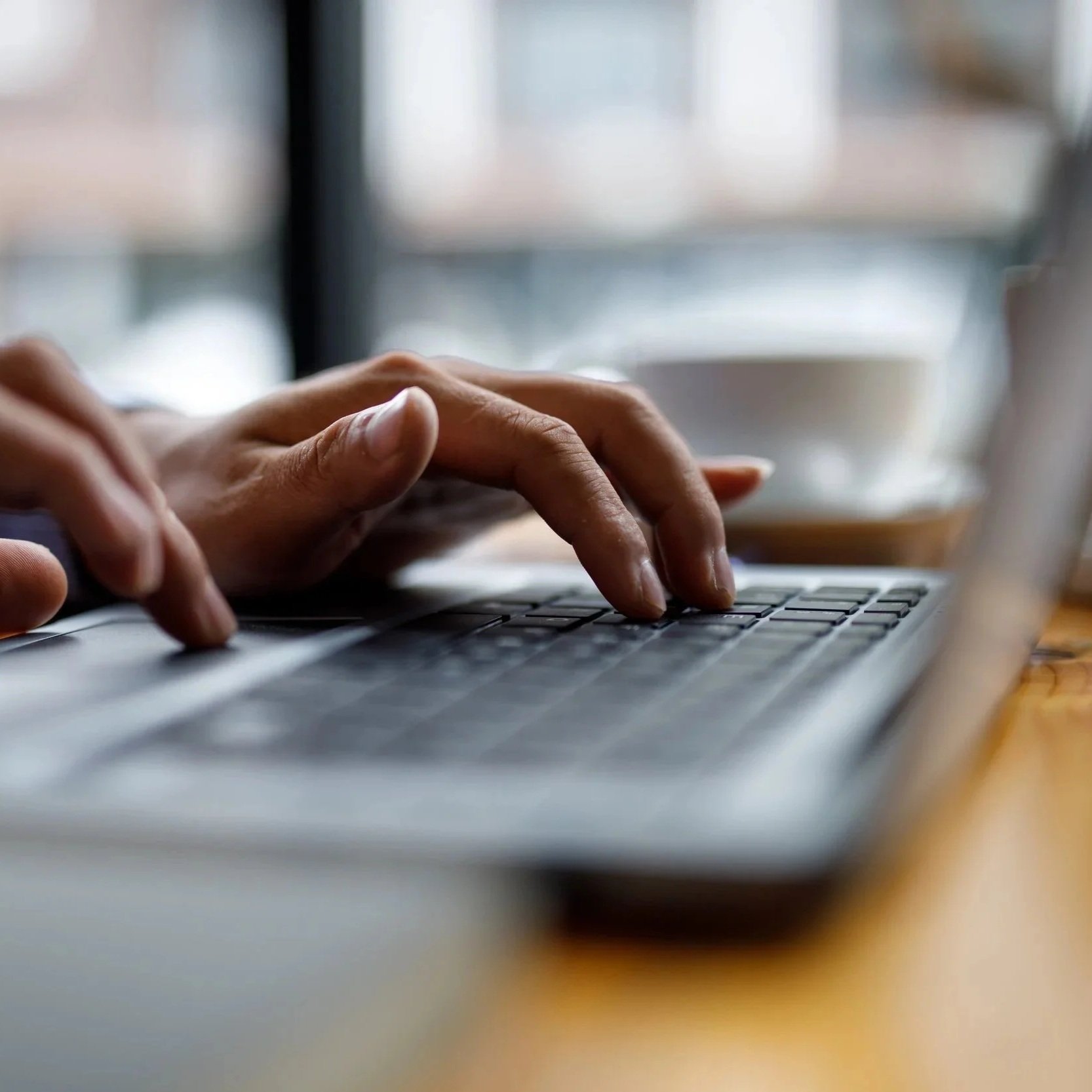 Close-up of hands typing on a laptop keyboard with a blurred background and a cup in the distance.