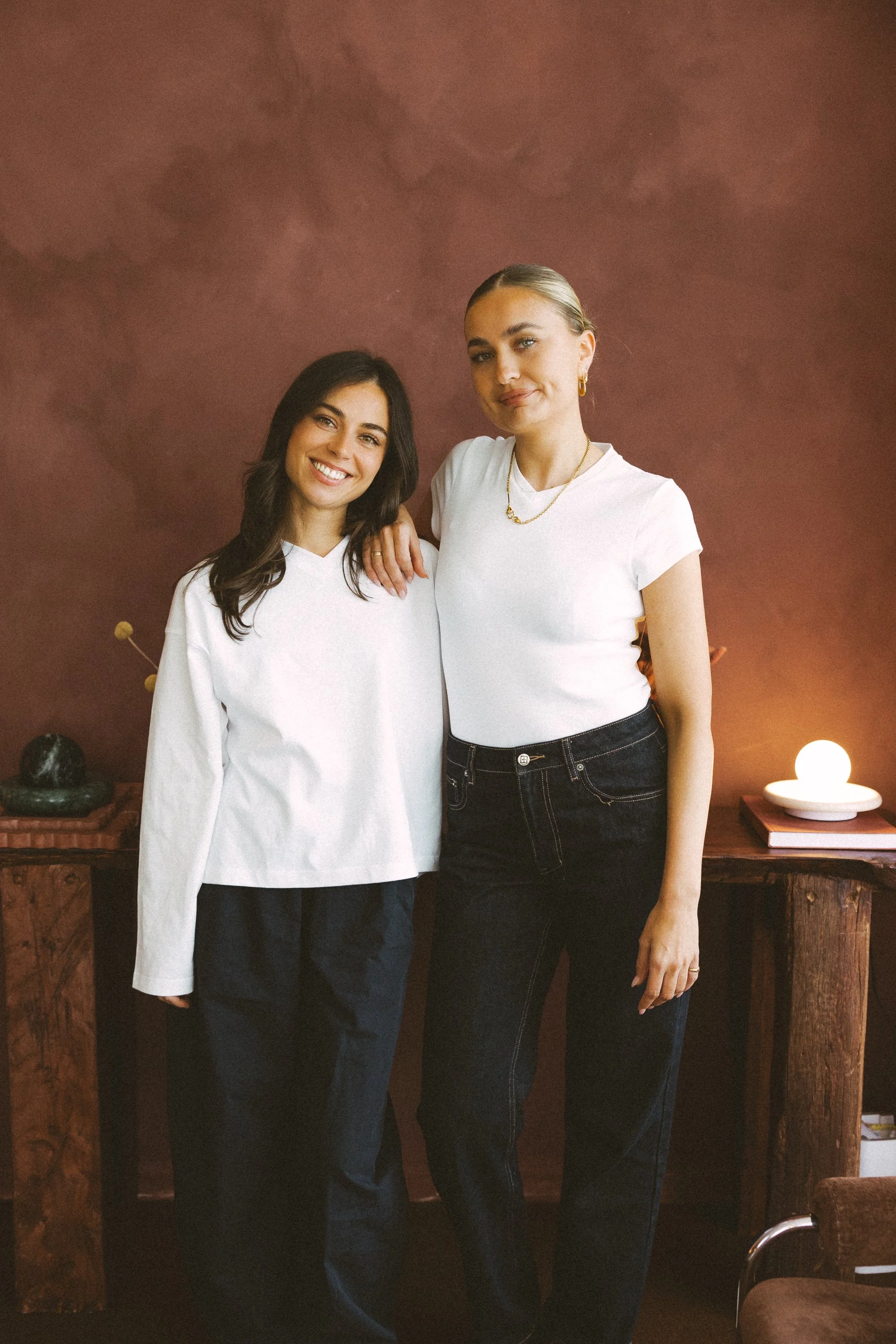 Two women standing close together and smiling, one with long dark hair and the other with blonde hair tied back, both wearing white tops and dark jeans, indoors with a wooden cabinet and warm lighting.