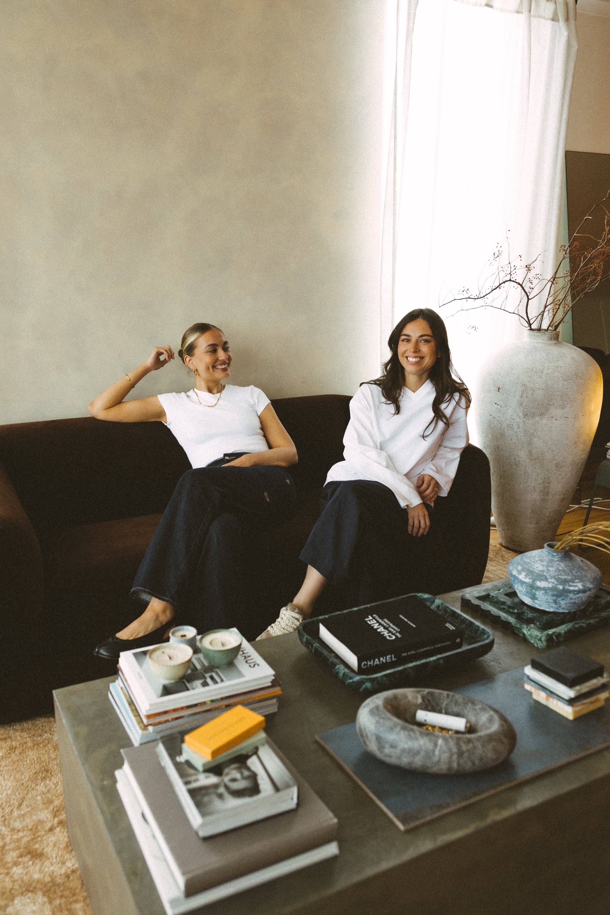 Two women sitting on a dark-colored sofa in a living room, smiling and talking. The woman on the left has blonde hair pulled back, wearing a white t-shirt, watch, and jeans. The woman on the right has dark, wavy hair, wearing a white hoodie, and black pants. A large ceramic vase with dry branches stands beside them. In the foreground, a low table holds books, magazines, candles, and decorative objects.