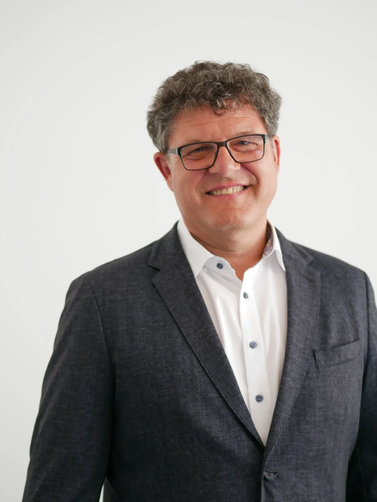 Ein lächelnder Mann mit Brille in einem dunklen Anzug vor weißem Hintergrund.