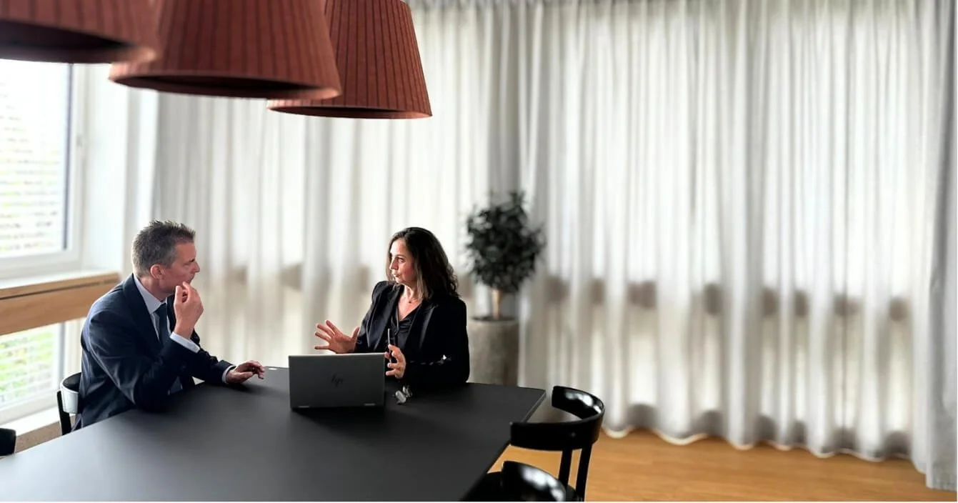 Zwei Menschen, ein Mann und eine Frau, sitzen an einem großen Konferenztisch und führen ein Gespräch, während ein Laptop geöffnet auf dem Tisch liegt. Der Raum ist hell erleuchtet mit großen Fenstern und weißen Vorhängen, und es gibt eine Topfpflanze im Hintergrund.