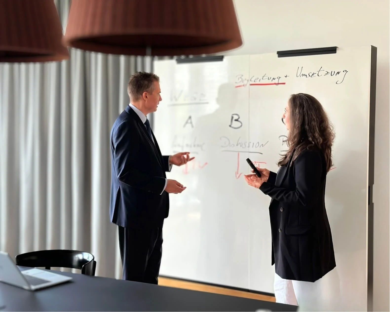 Ein Mann und eine Frau unterhalten sich vor einer weißen Tafel mit handschriftlichen Notizen in einem Büro.