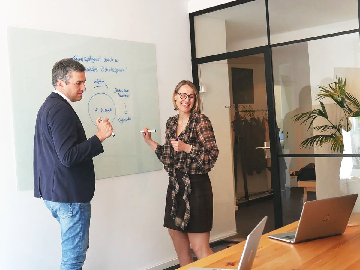 Zwei Personen, ein Mann und eine Frau, arbeiten zusammen in einem modernen Büro, der Mann schreibt auf eine Whiteboard mit Notizen und der Frau lacht. Ein Tisch mit Laptops ist im Vordergrund.