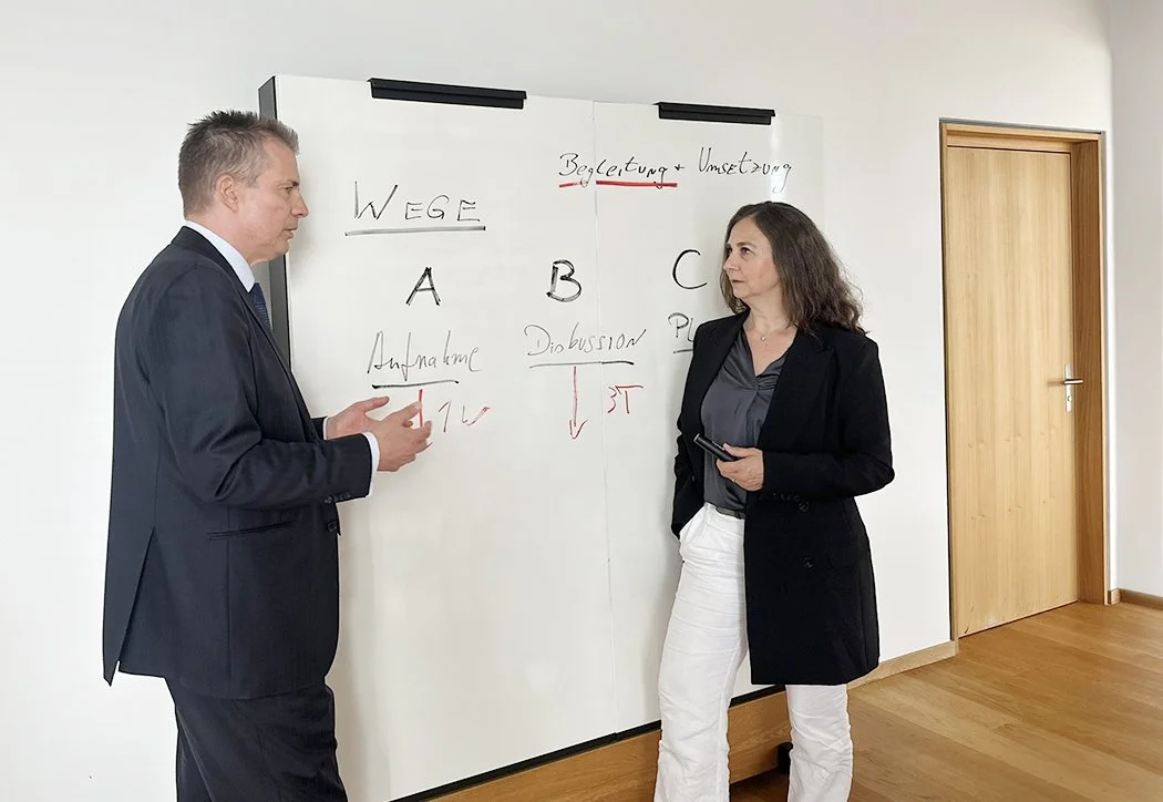 Zwei Personen, ein Mann und eine Frau, stehen vor einem Whiteboard und führen eine Diskussion. Das Whiteboard enthält handgeschriebene Begriffe und Diagramme, darunter 'WEGE', 'A', 'B', 'C', 'Begleitung + Umsetzung', 'Aufnahme', 'Discussion' und verschiedene Pfeile.