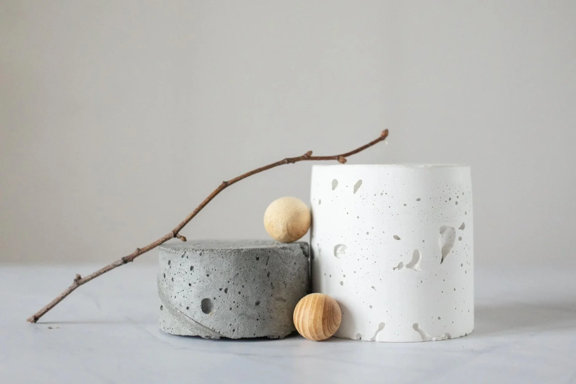 Minimalist arrangement with two concrete and ceramic vessels, a dried twig, and wooden spheres on a white surface against a plain background.