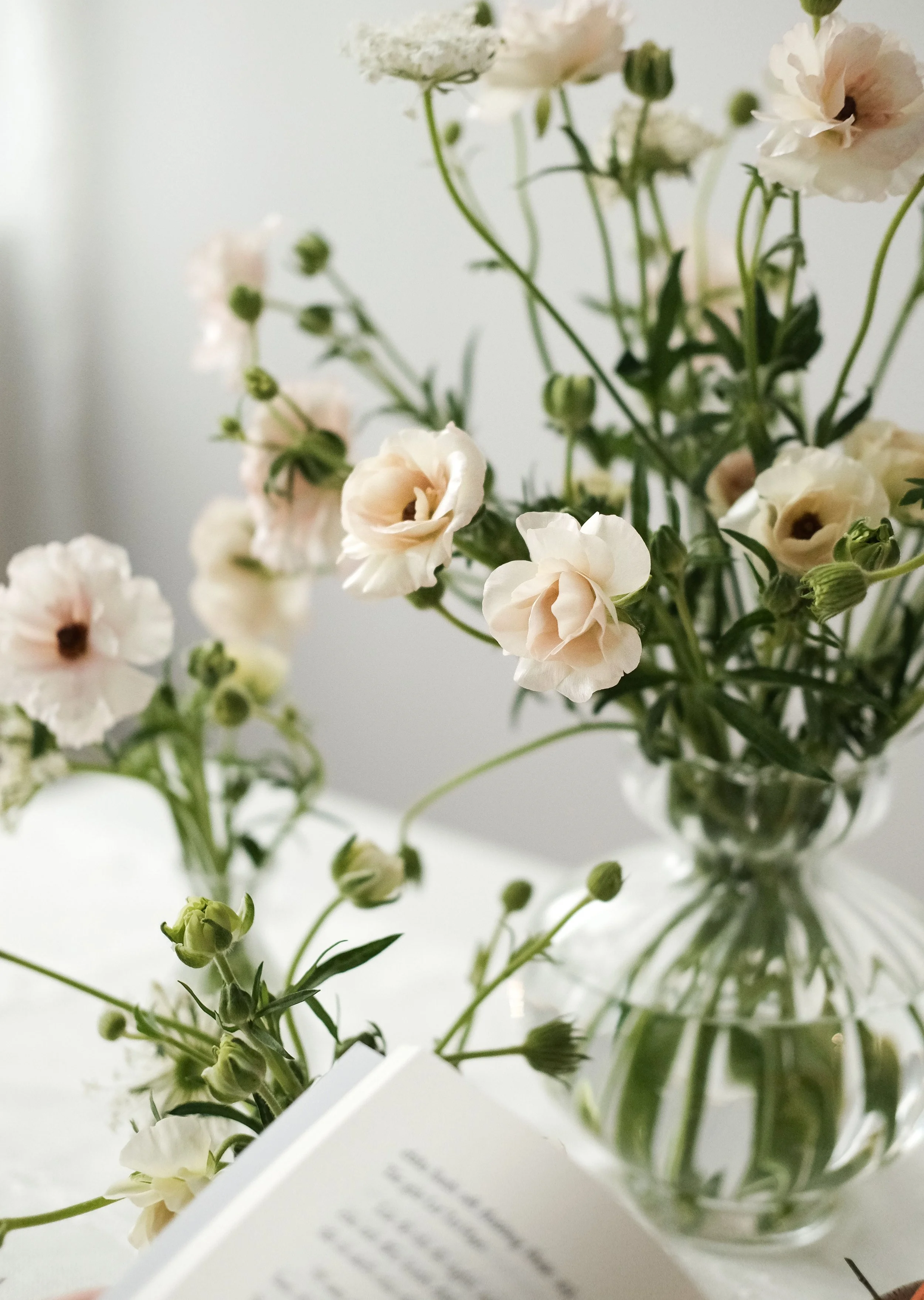 Close-up of a bouquet of white and pale pink flowers, possibly ranunculus, in a glass vase with some greenery.