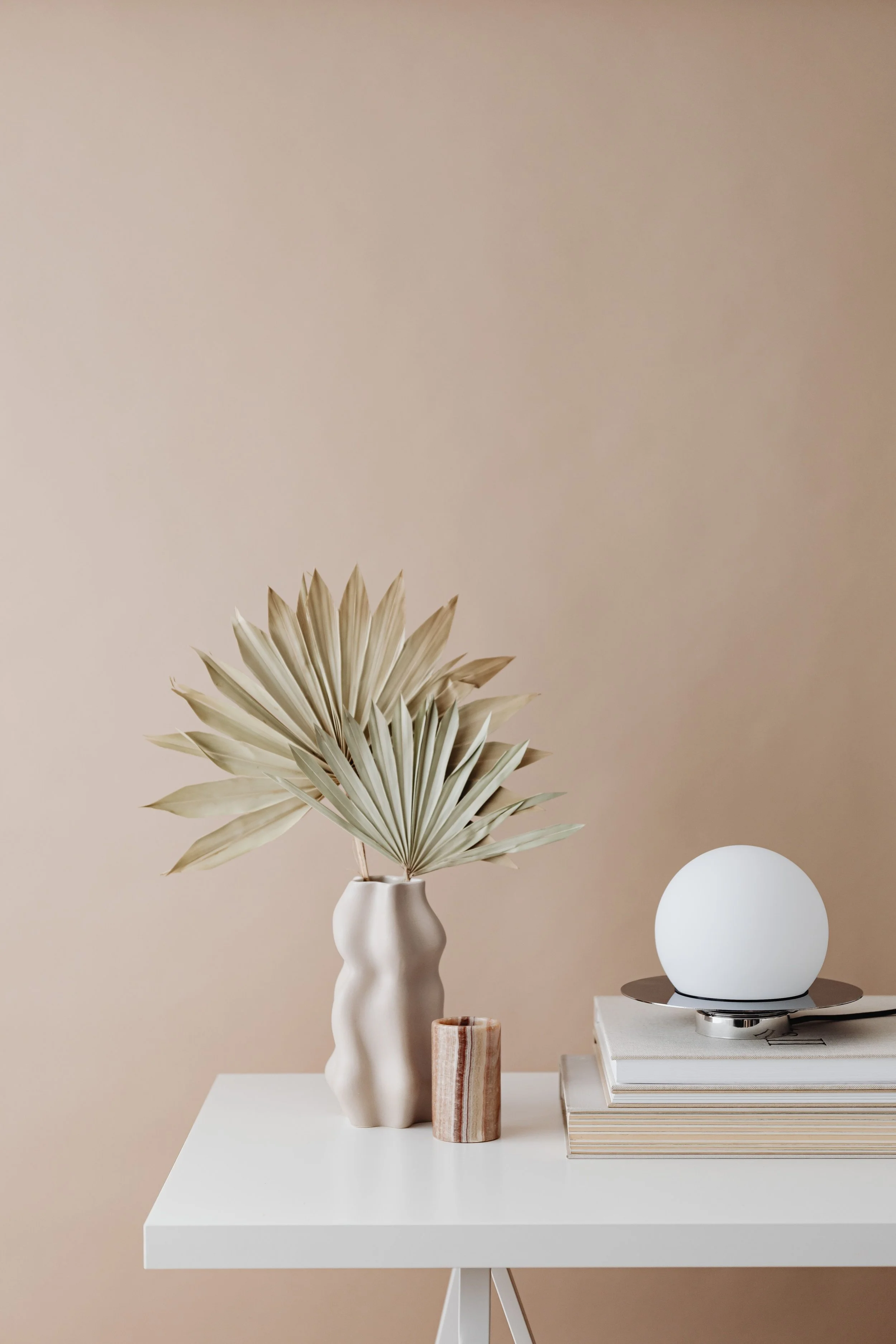 Minimalist living space decor with a beige wall, a white vase with large dried palm leaves, a small marble-patterned decorative item, a white spherical lamp, and a stack of books on a white table.