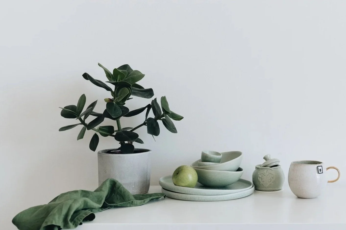 A potted green leafy plant, a green apple, stacked white ceramic bowls, a small decorative jar, and a white mug with a handle on a white tabletop against a plain light-colored wall.