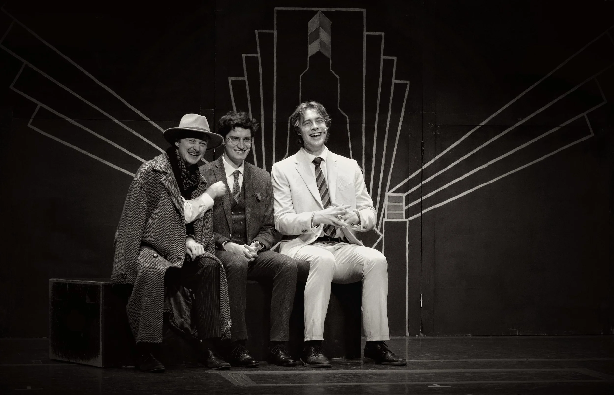 Three men and one woman sitting on a bench, smiling and laughing on a stage with a dark background and a geometric design outline.