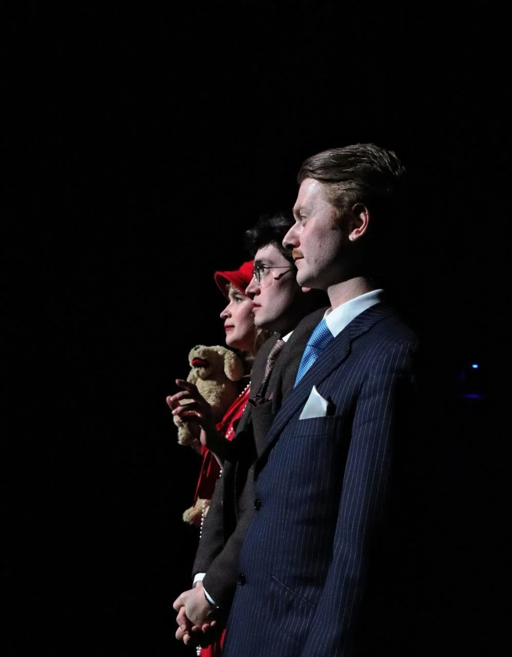Side profile of three individuals standing in line on a dark stage, dressed in vintage attire, with serious expressions. The woman is holding a teddy bear.