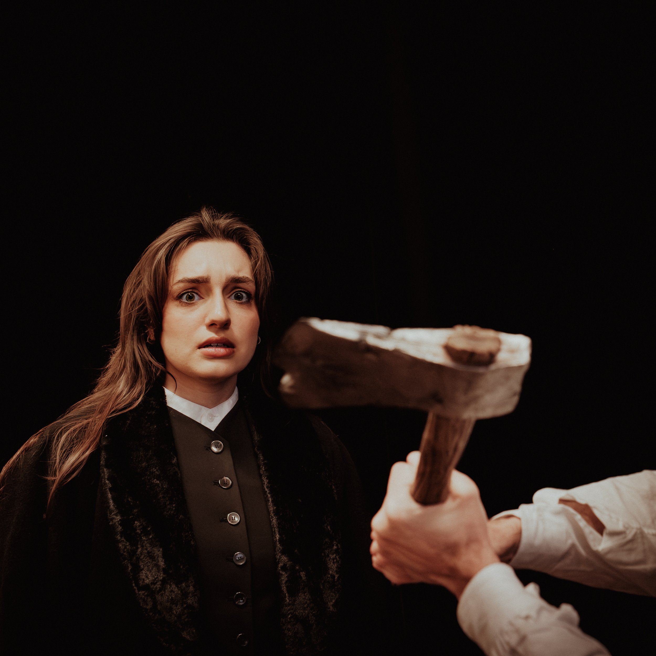 A woman with light brown hair, wearing a black shirt with buttons and a black jacket, looking surprised or curious, standing in front of a large skeleton held by another person with long reddish hair, all set against a dark background.