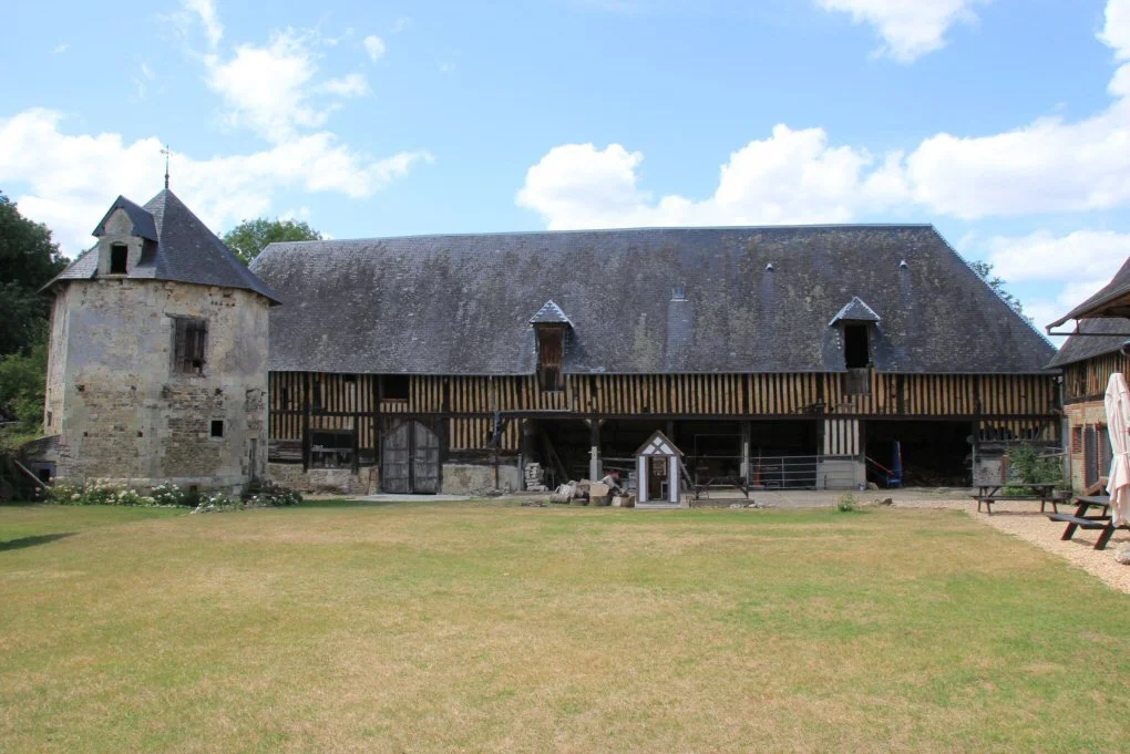 An old stone building with a pointed roof and a wooden barn with a slate roof, set against a partly cloudy sky. There is a grassy yard in front with a few benches and a small statue.