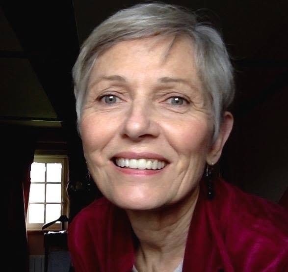 A smiling middle-aged woman with short gray hair, wearing a red top, indoors with a window in the background.