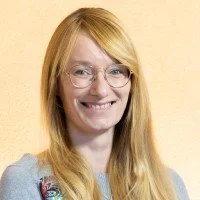 A young woman with long blonde hair, glasses, and a big smile, standing against a light-colored background.