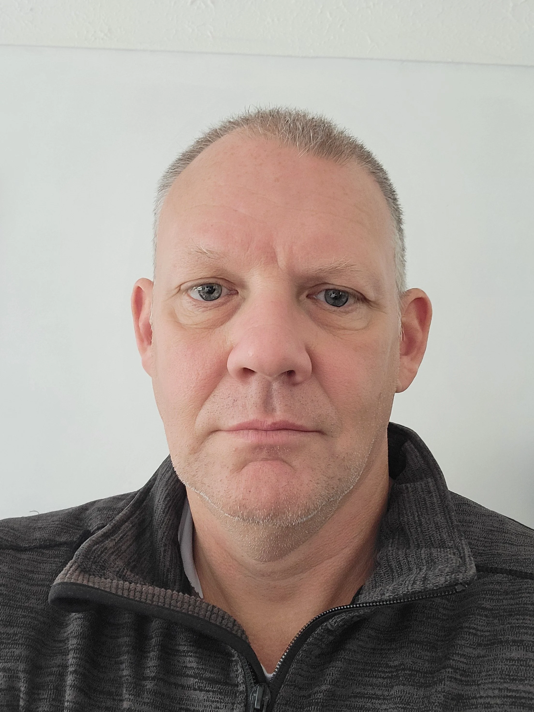 Close-up of a middle-aged man with short hair and blue eyes, wearing a black zip-up jacket, against a plain white wall.