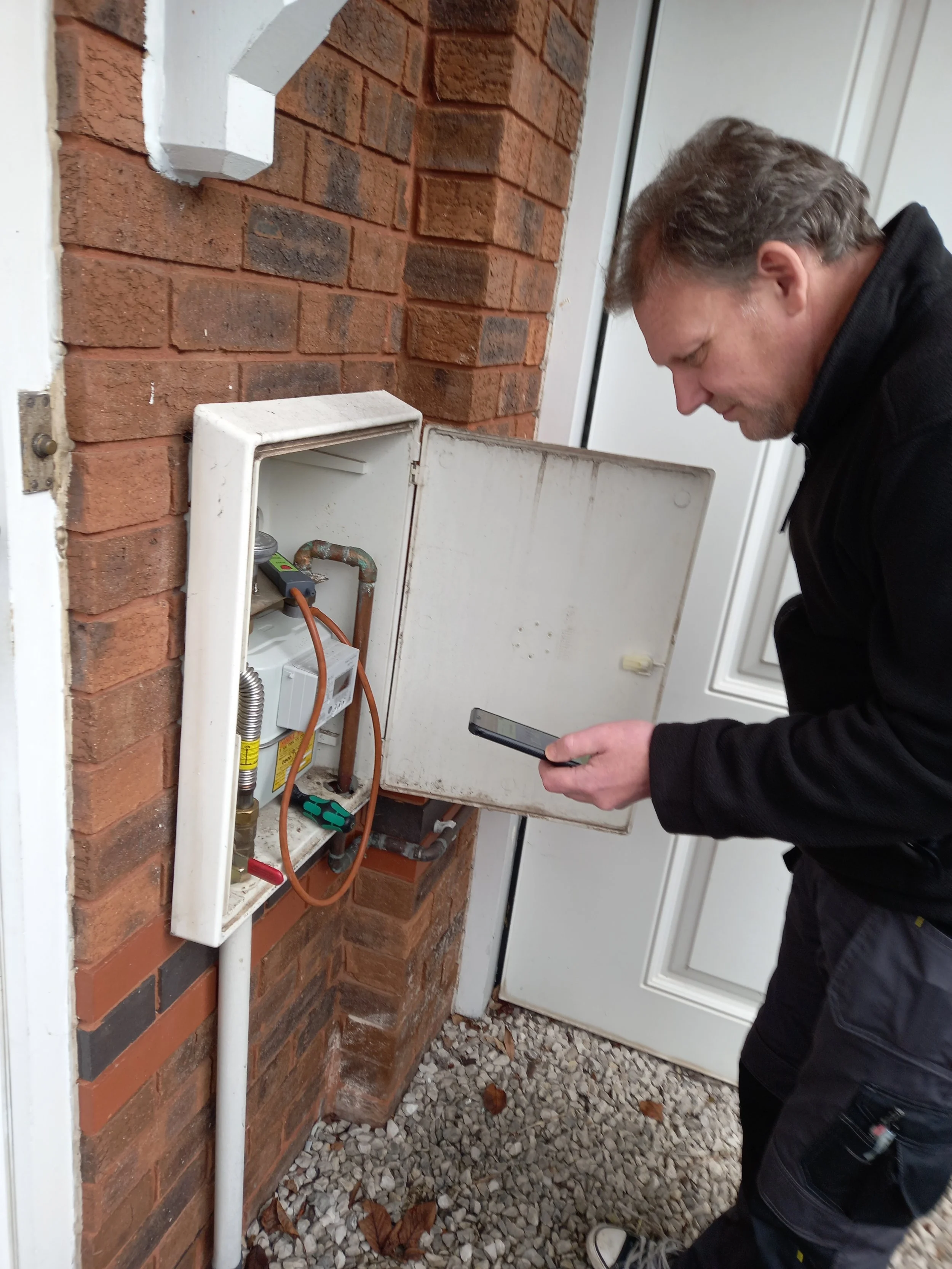 A man inspecting an open electrical box mounted on a brick wall outside a house.