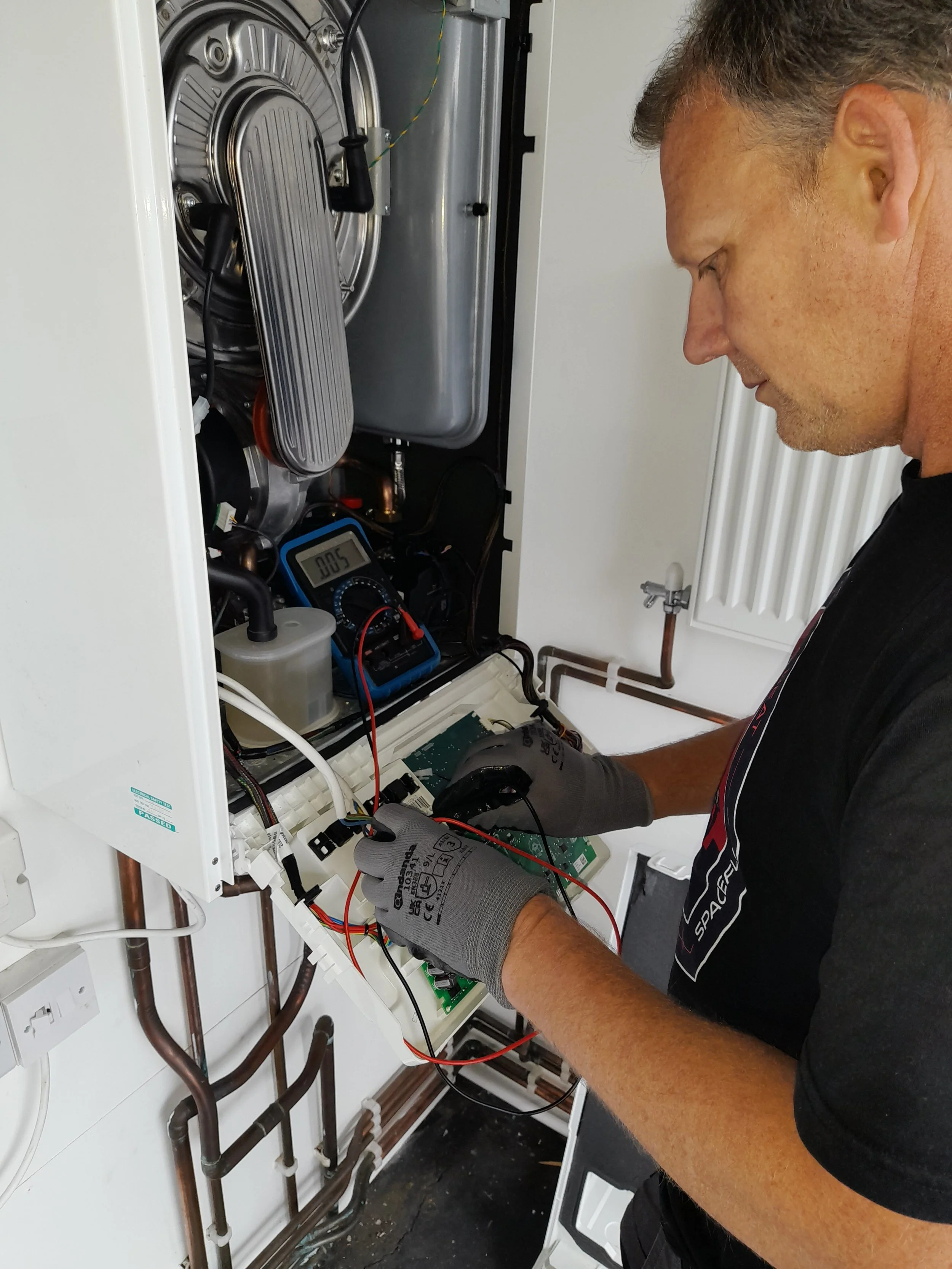 A technician is repairing or inspecting an appliance, possibly a boiler or heating system, inside a white cabinet. The technician is wearing gloves and is using a multimeter or similar device to test electrical connections.