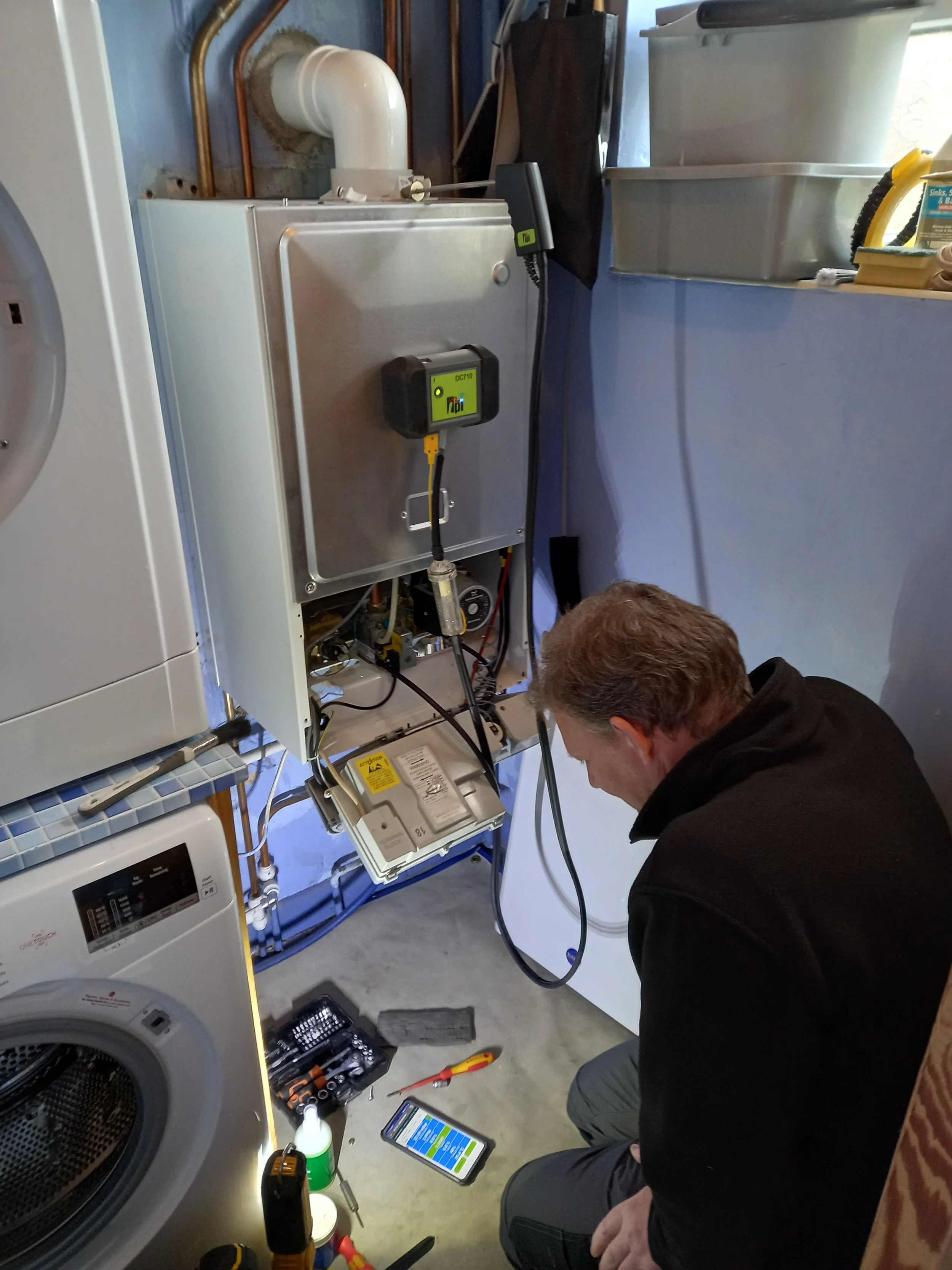 A person is working on appliance repair in a laundry room. He is crouched on the floor, using tools with a toolbox nearby. Wires, parts, and a smartphone are also on the floor. The repair appears to involve a metallic electric or gas appliance, possibly a dryer or washing machine, with various tools and electronic devices around.
