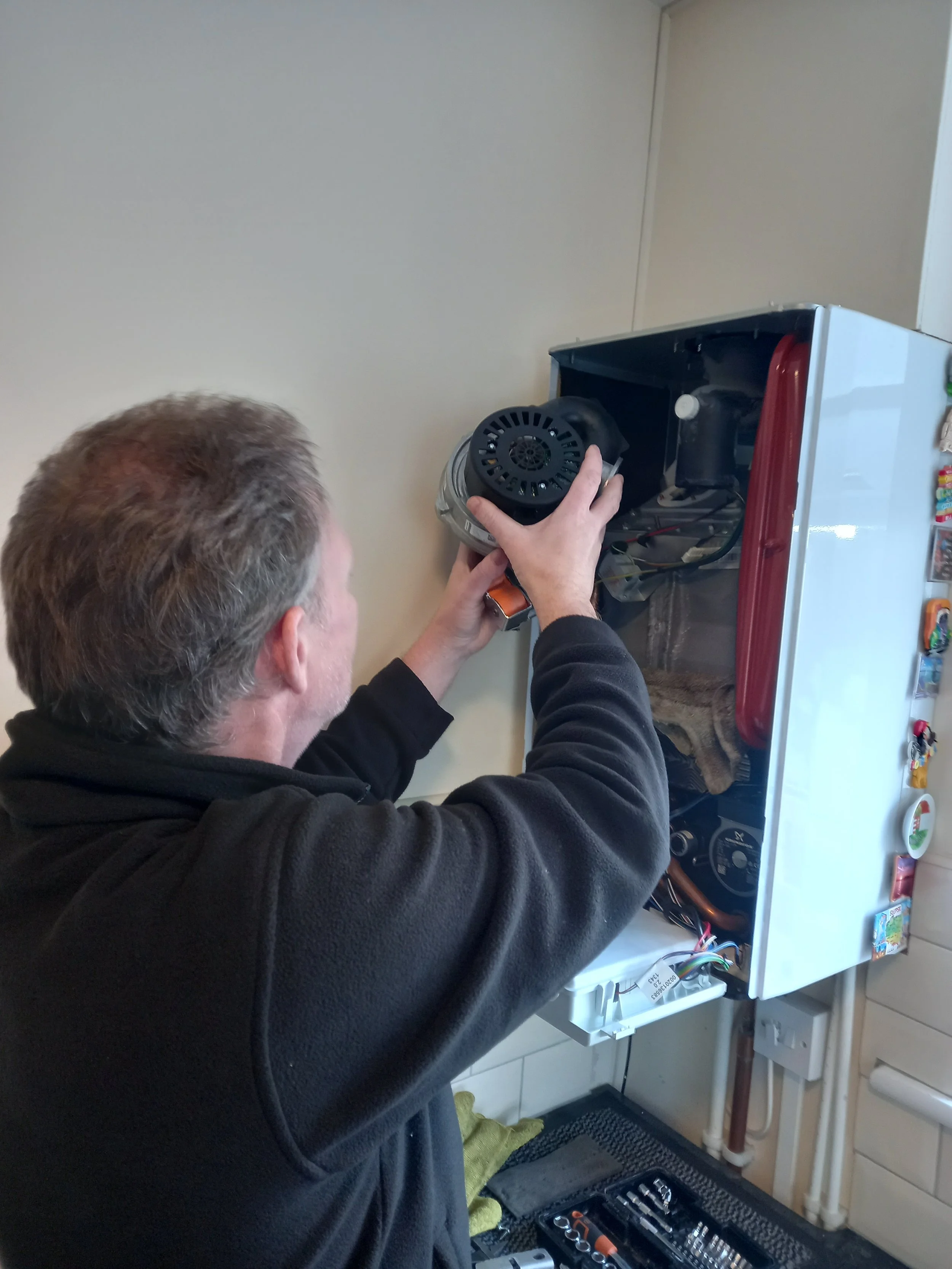 A man working on an appliance, possibly a refrigerator, with an open back panel. He is holding part of the appliance with one hand and wearing a black jacket. Tools are laid out on a mat on the floor nearby, and the setting appears to be a kitchen with a white wall and some shelves.