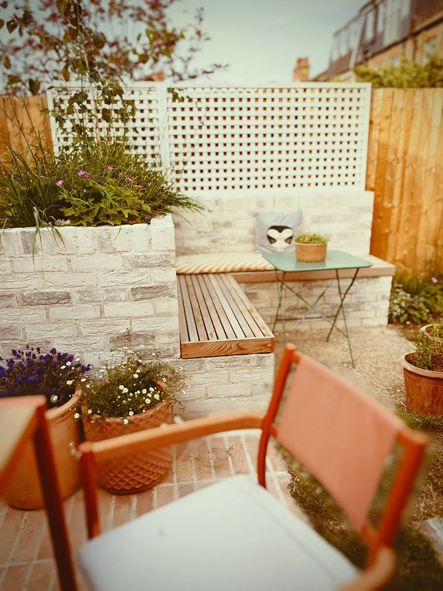 A cozy outdoor patio area with a brick bench, potted plants, a small turquoise table, and a white lattice privacy screen, surrounded by a wooden fence and flowering plants.