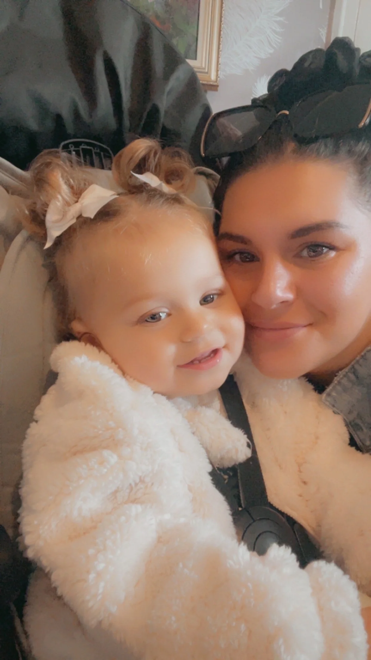 A woman and a young girl taking a selfie together, both smiling and close to each other. The girl has her hair in two buns tied with white bows, and the woman has dark hair styled with a large black bow.