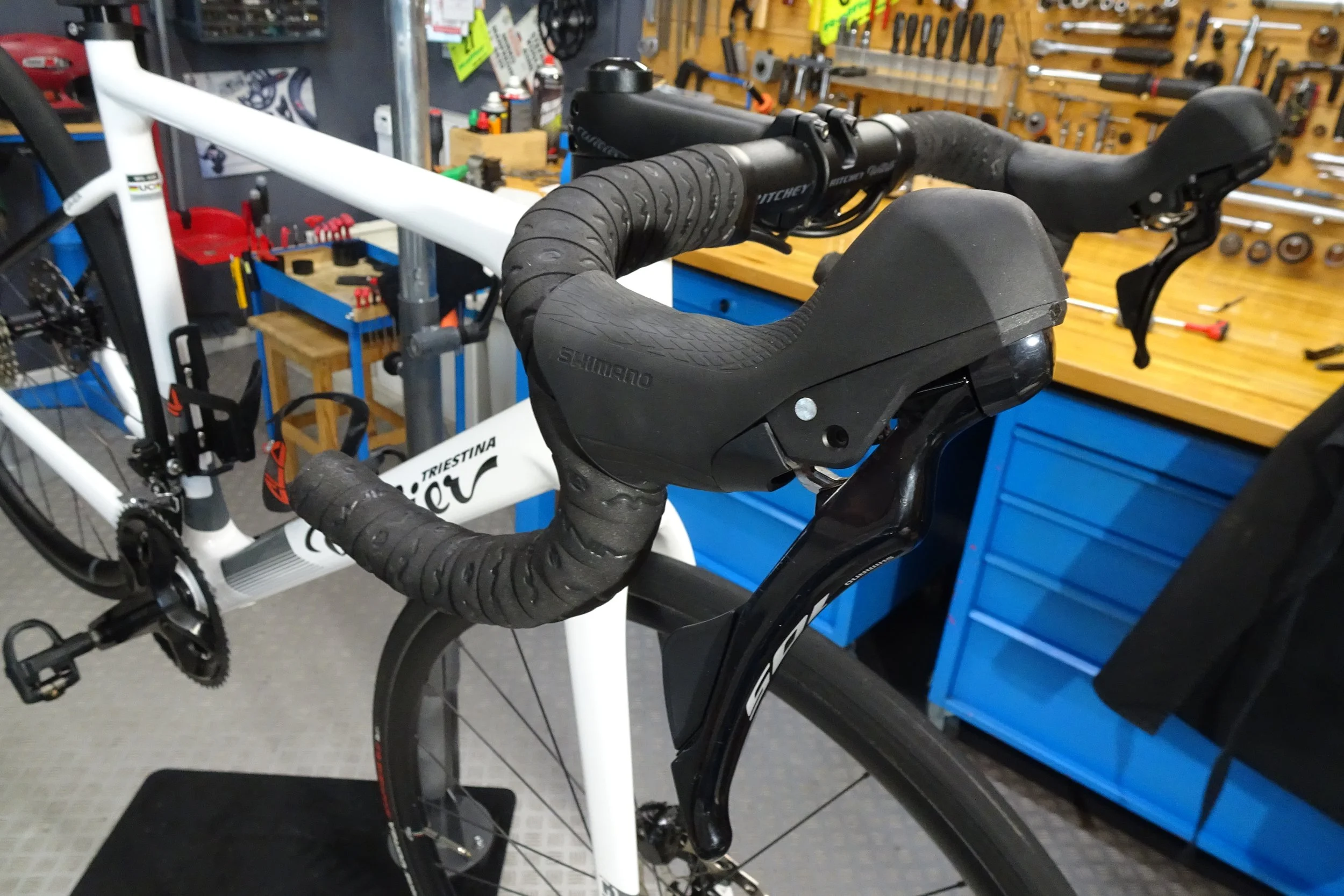 Bici da corsa con manubrio a scafo nero e bianco, all'interno di un'officina con pareti attrezzate con attrezzi da ferramenta.