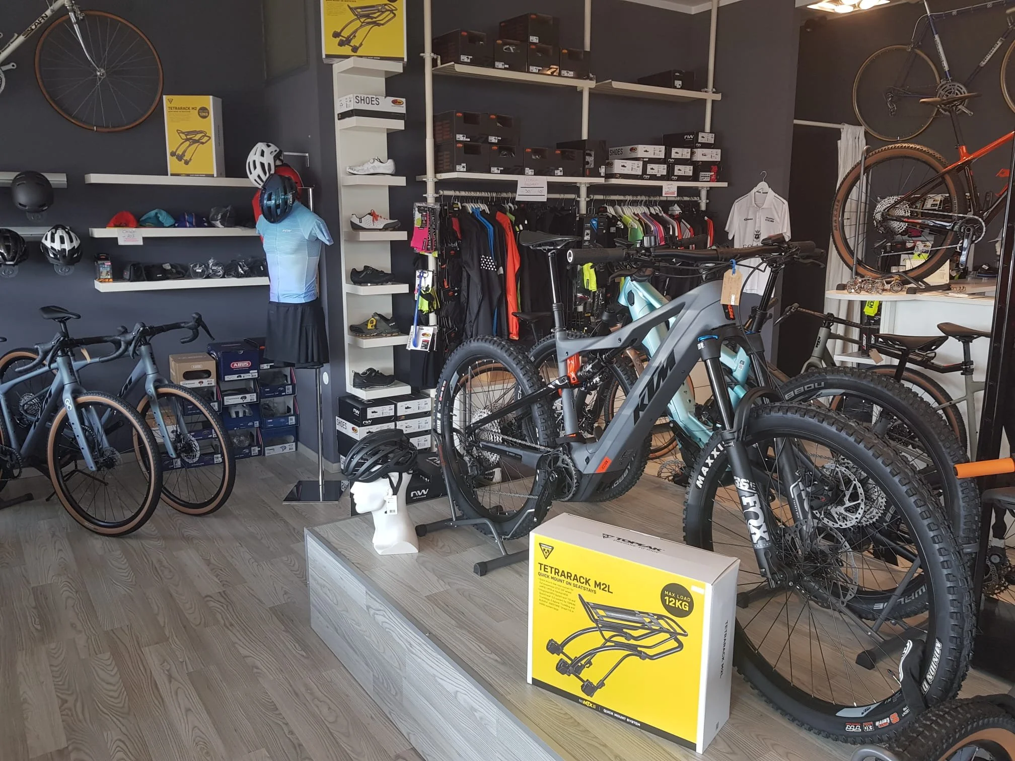 Interno di un negozio di biciclette con biciclette, caschi e accessori esposti, scaffali con scatole e vestiti sportivi, motivo di sfondo scuro e pavimento in legno.