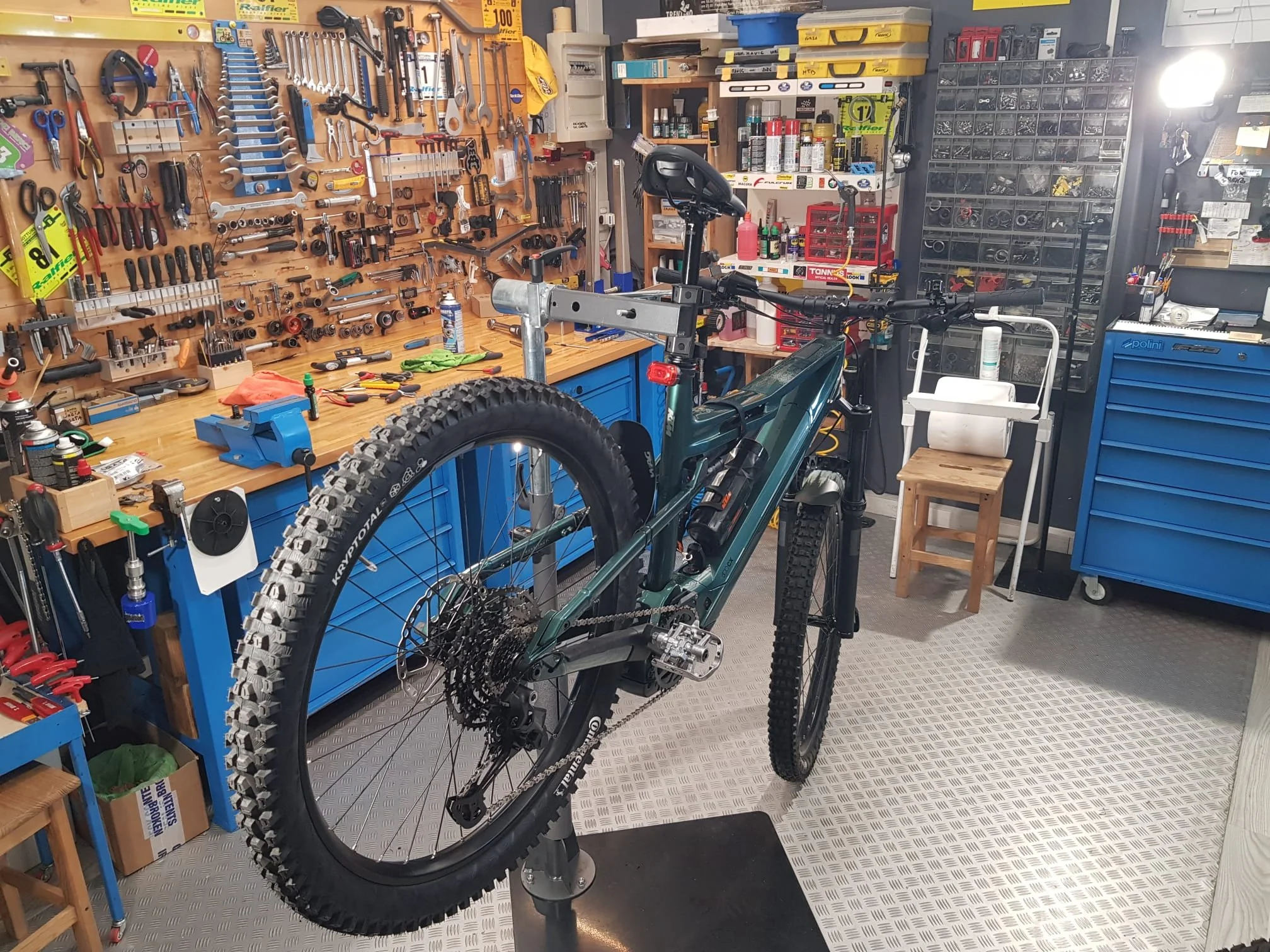 Un'officina di riparazione bici con attrezzi appesi alle pareti, scaffali con materiali e un cicli in fase di riparazione su un supporto.