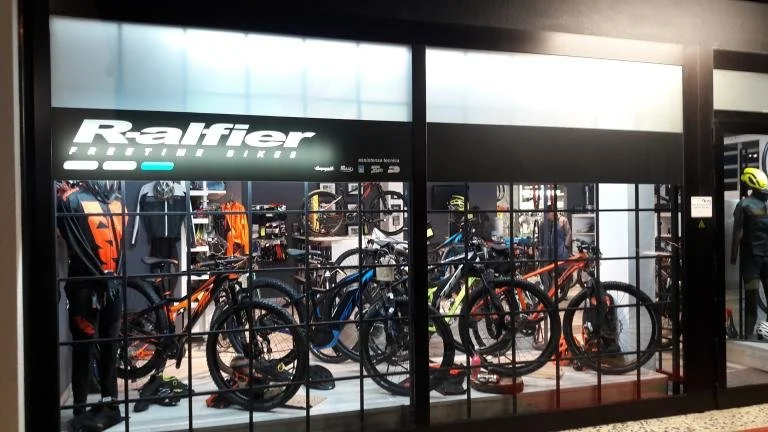 Display window of a bicycle store named 'Ralfier', showing mountain bikes, cycling gear, mannequins dressed in cycling apparel, and helmets.