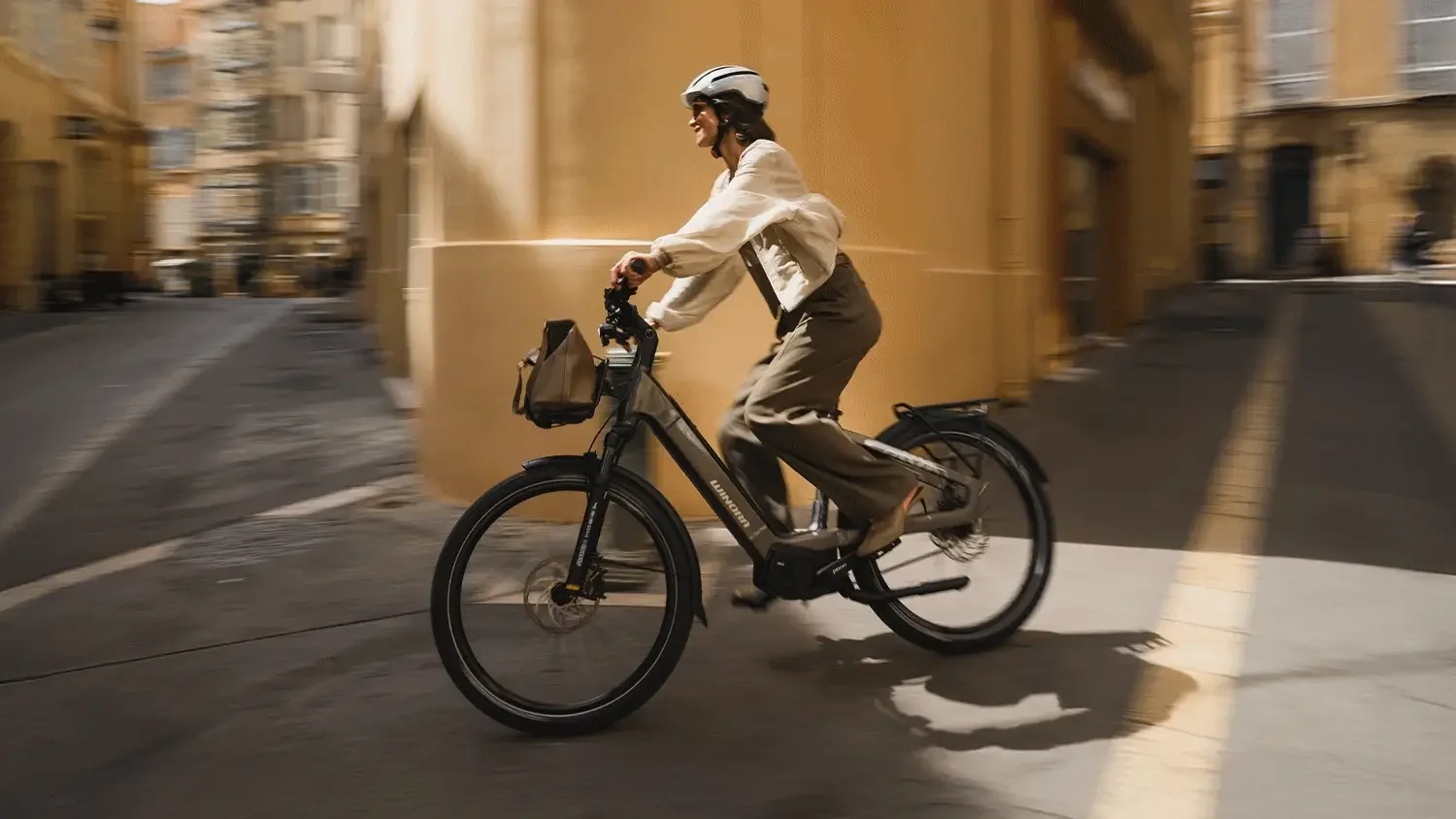 Una donna felice che pedala in bicicletta in una strada urbana. Indossa un casco e occhiali, con una borsa attaccata al manubrio, sfondo di edifici gialli.