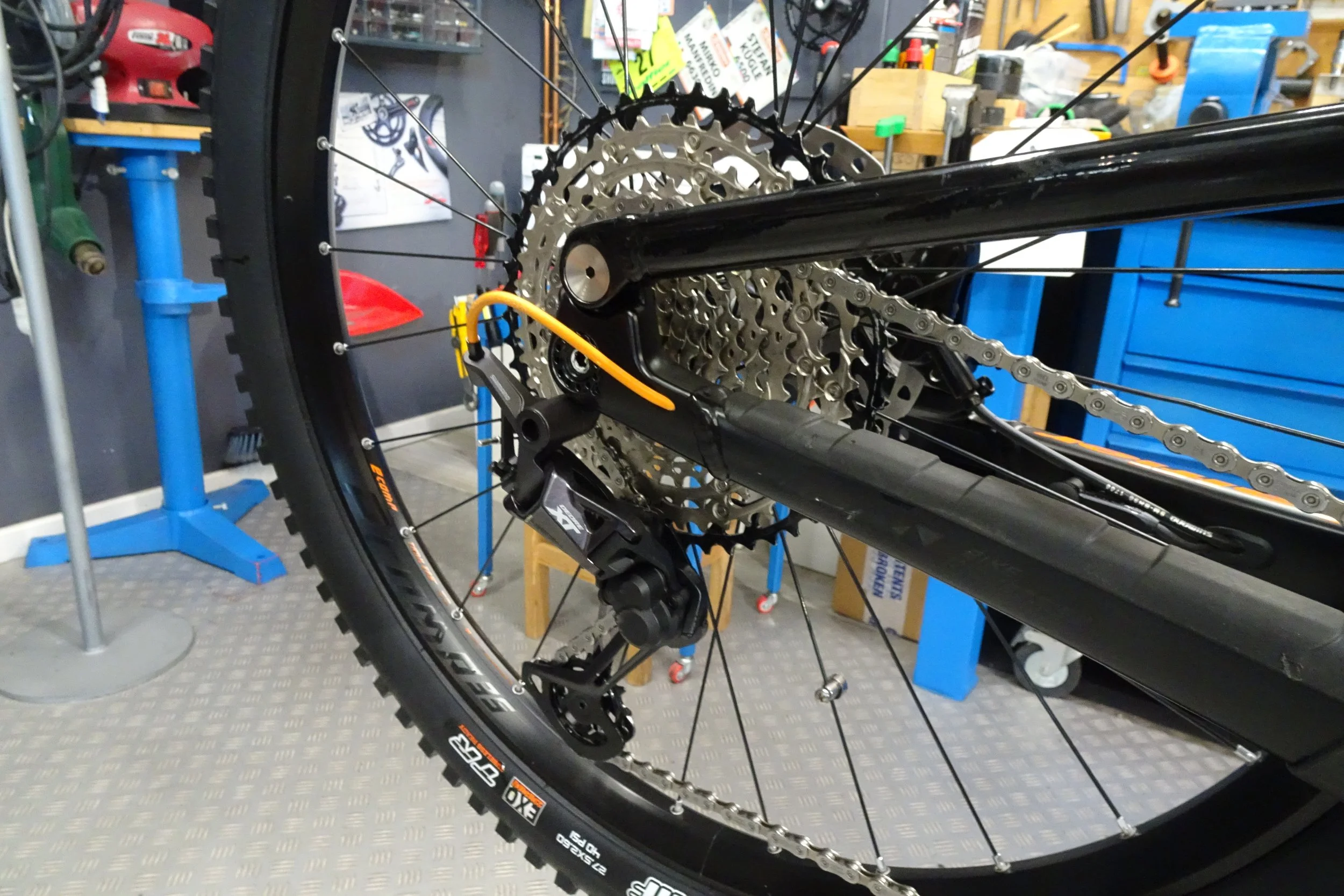 Particolare di una bicicletta da mountain bike in officina, focale sul sistema di trasmissione con vari pignoni e catena, con attrezzi e strumenti di lavoro sullo sfondo.