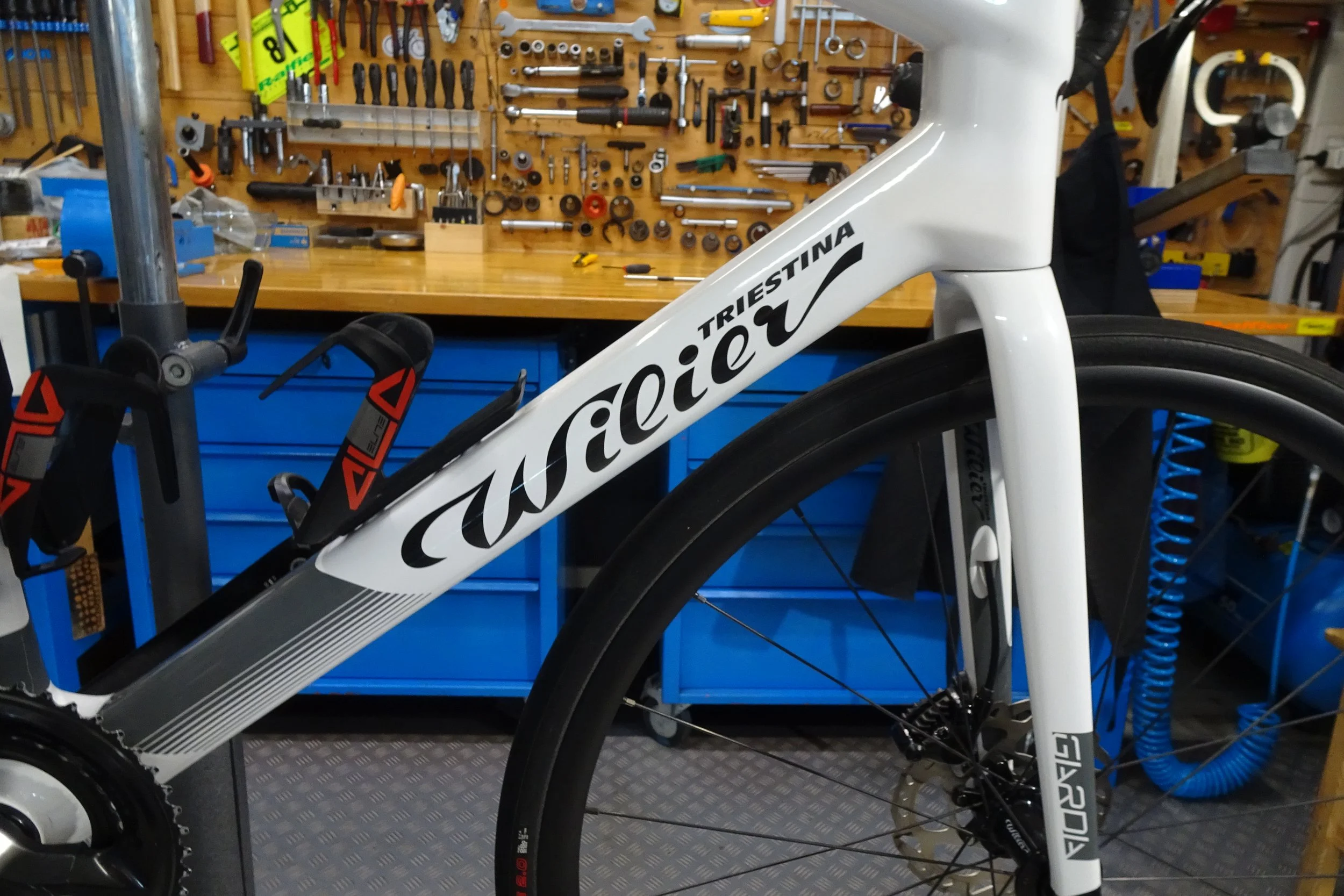 Una bicicletta bianca con la scritta 'Triestina' e 'Wilier' sul telaio si trova in un'officina, con un banco da lavoro e attrezzi appesi sullo sfondo.