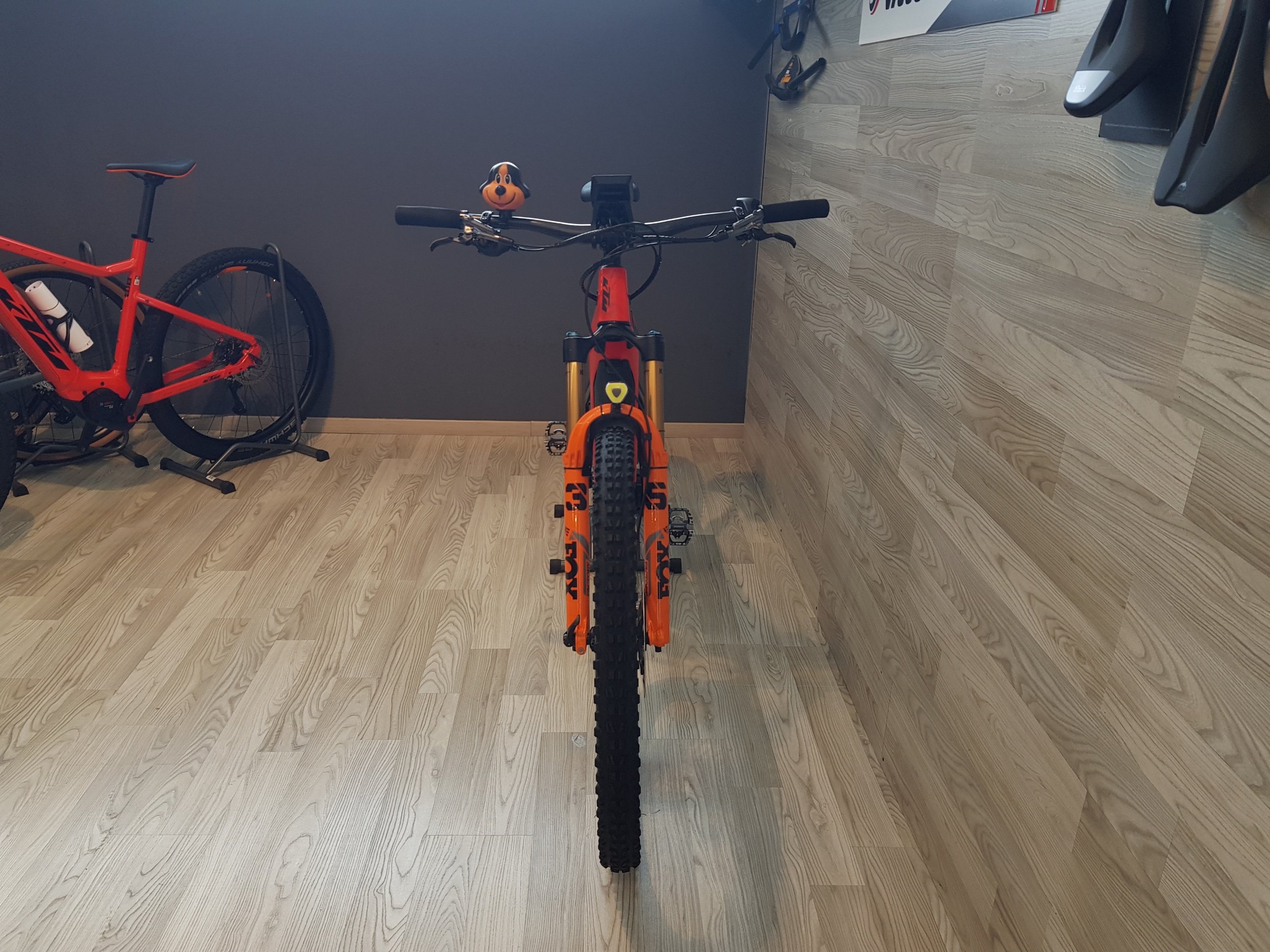 Bici da montagna arancione e nera con manubrio, davanti a una parete grigia chiara, sul pavimento in legno.