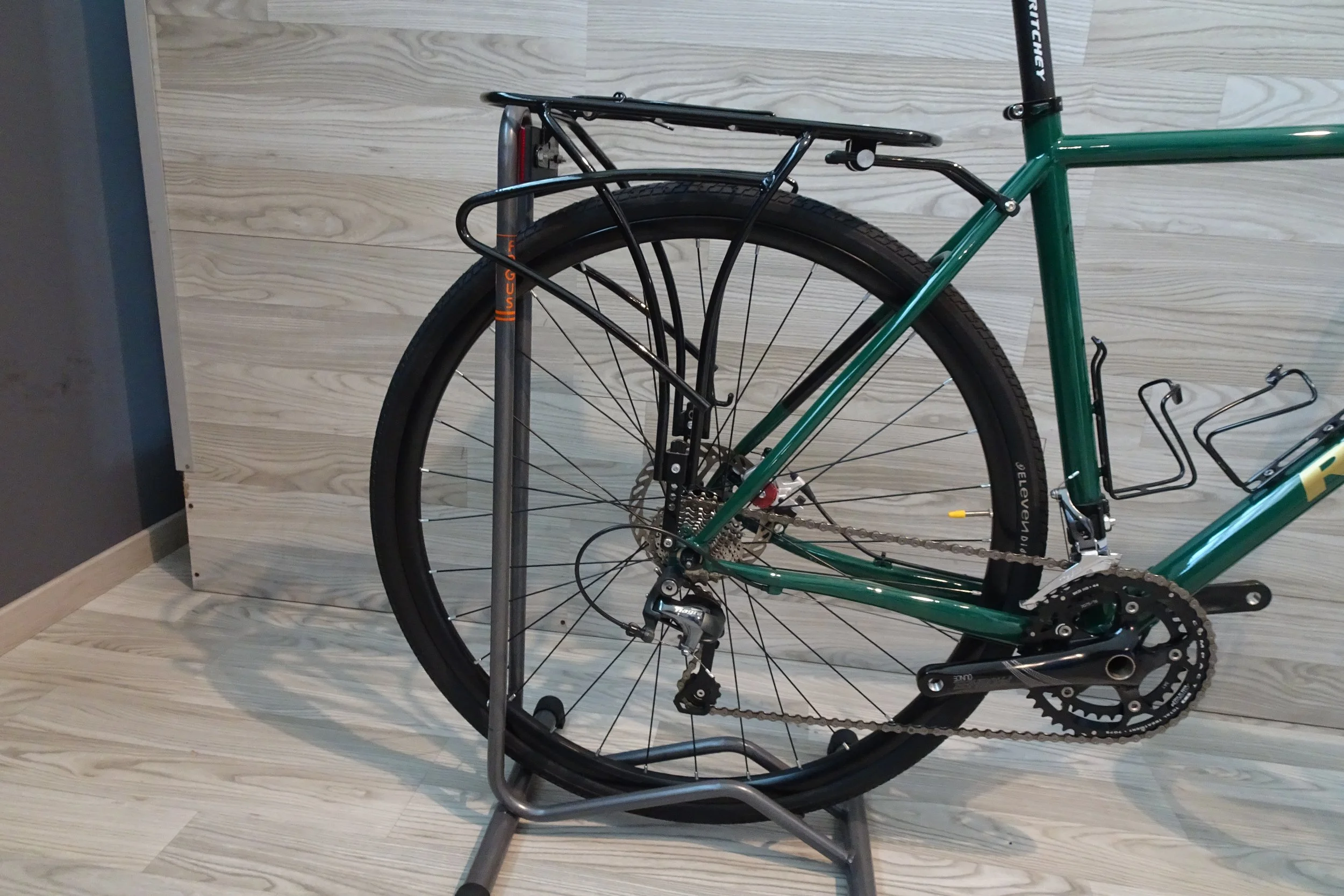 Bicicletta verde con portapacchi posteriore, appoggiata su un supporto in metallo, con sfondo di parete di legno chiaro.