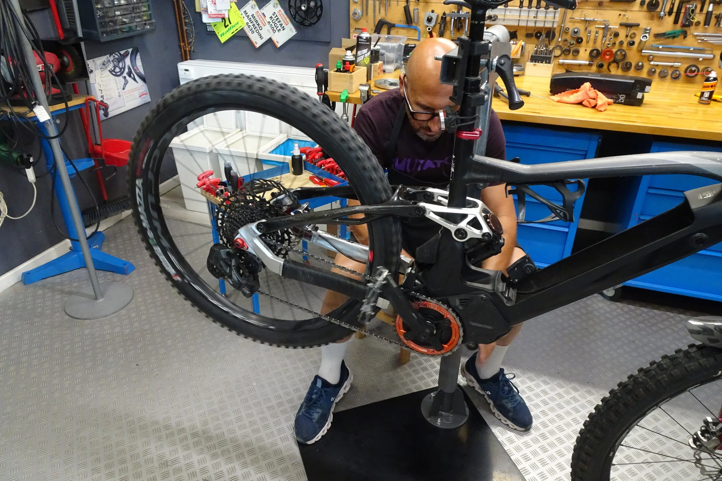 Un uomo con occhiali e lampada frontale sta lavorando su una bicicletta in un laboratorio attrezzato con attrezzi e scaffali di vario tipo.