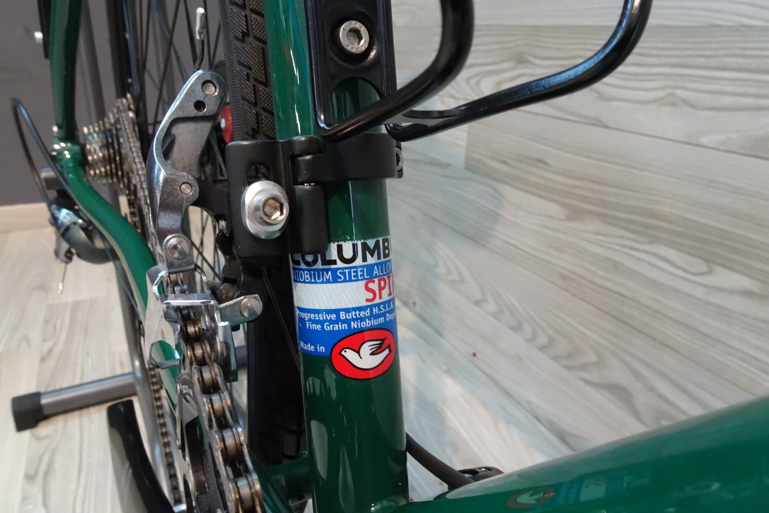 Immagine di una bicicletta vista frontalmente con il telaio verde, la catena, i pignoni e i deragliatori. Sul tubo del telaio è presente una etichetta con il logo di una colomba bianca.