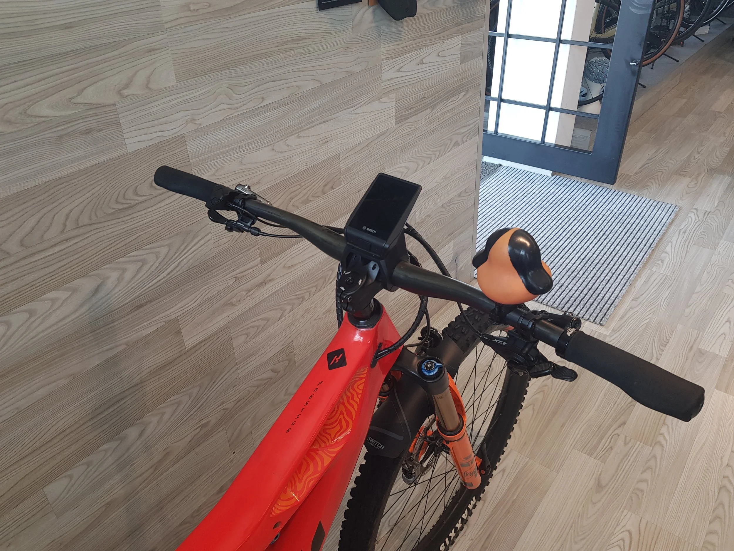 Bicicletta da mountain bike rossa con manubrio e sella, vista dall'alto in un ambiente indoor con pavimento in legno e un ingresso o uscita con vetro e metallo.